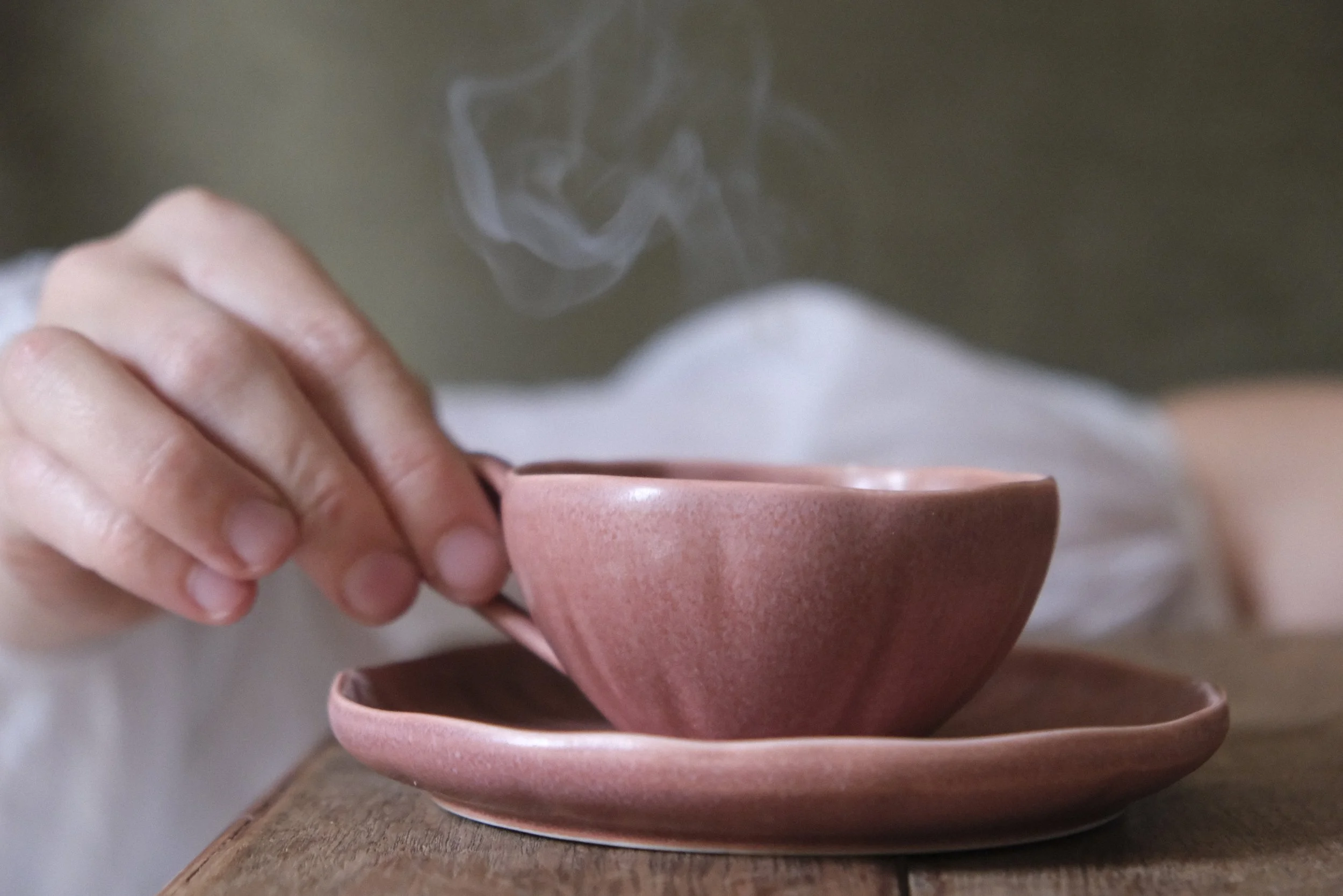 Close-up of a hand holding a steaming cup of tea or coffee, with a second cup underneath on a wooden surface.
