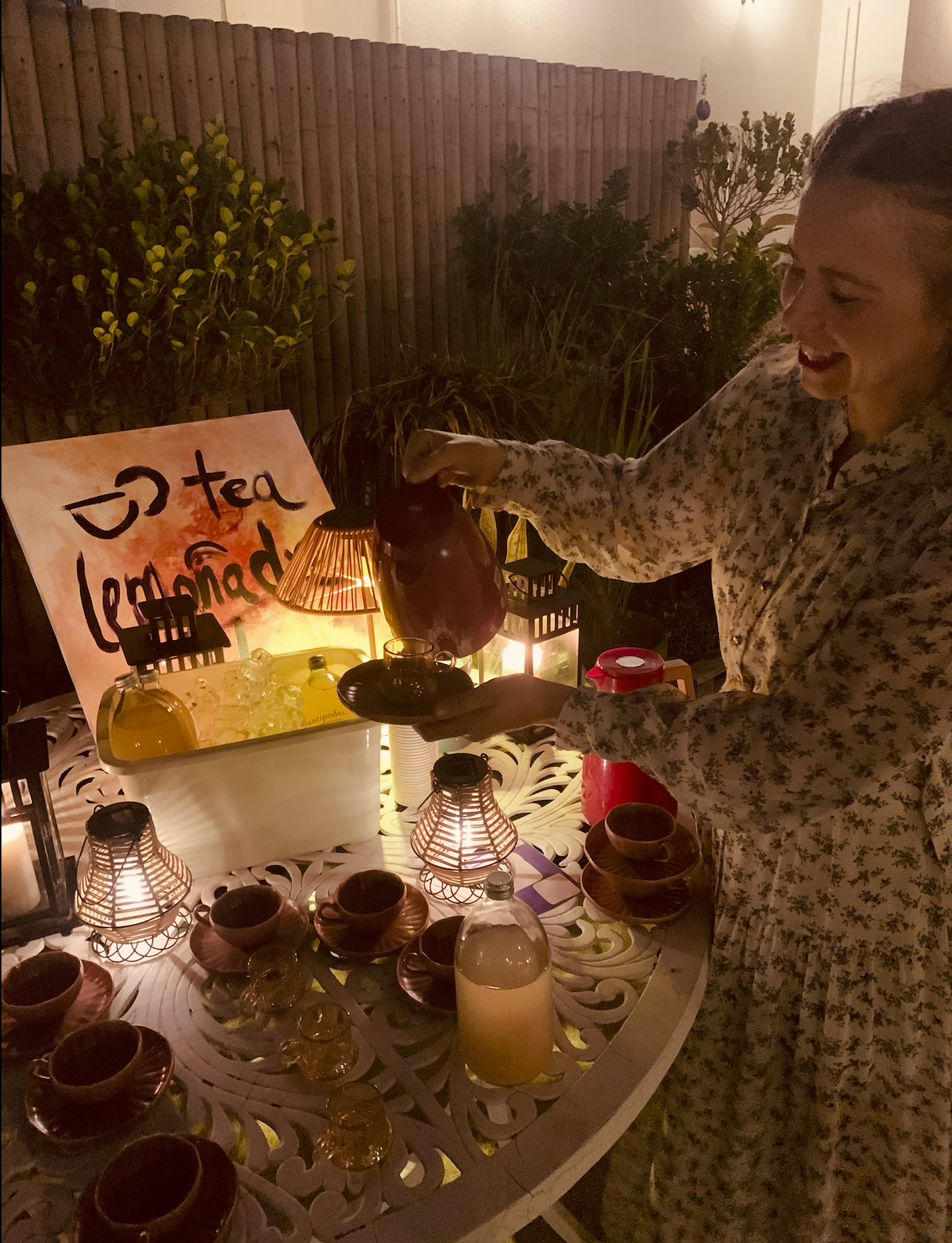 A woman in floral pajamas preparing tea at an outdoor evening tea party with a sign that says "Tea Lemonade," a large container of lemonade, several tea cups, and decorative lanterns on the table.