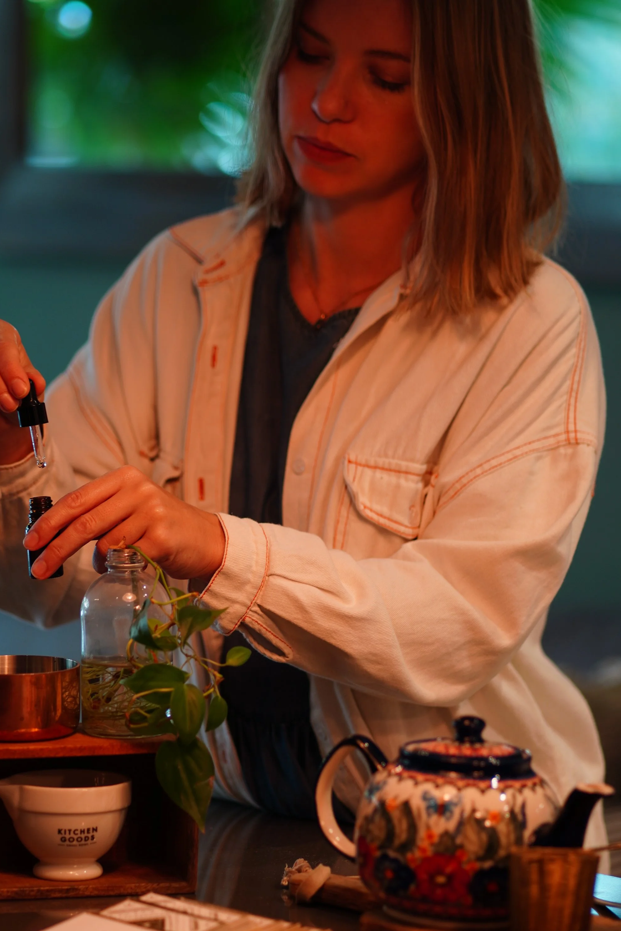 A woman with shoulder-length hair wearing a beige jacket and dark shirt preparing a drink at a kitchen counter. There are various kitchen items including a floral patterned teapot, a small plant, cups, and utensils on the counter.