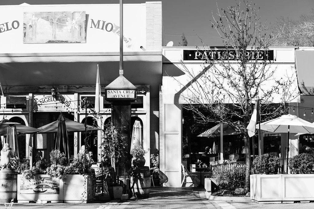 Black and white photo of a street scene with restaurants, outdoor seating, umbrellas, potted plants, a leafless tree, and building facades with signs and large windows.