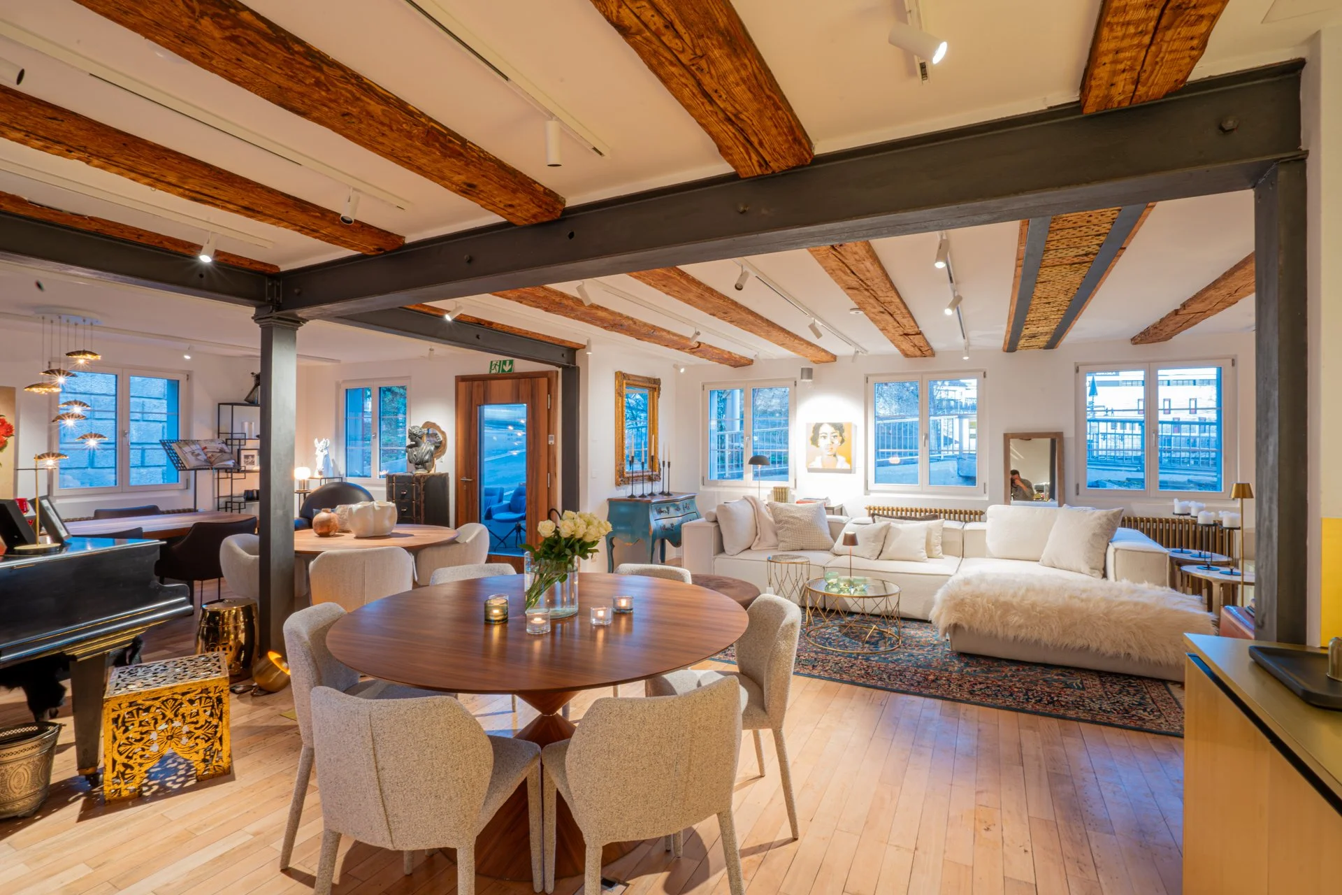 Innenansicht eines modern eingerichteten Wohnzimmers mit Holzbalkendecke, großen Fenstern, einem weißen Sofa, einem runden Holztisch mit Stühlen und Dekorationsobjekten.