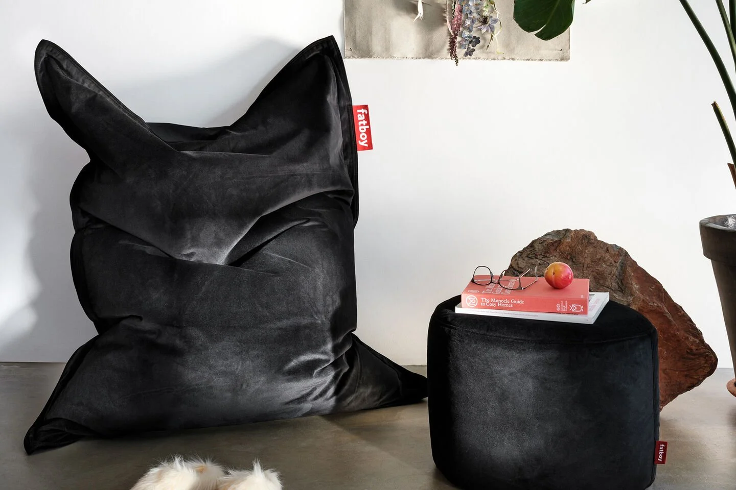 Ein schwarzer Sitzsack, eine schwarze Hocker, ein rotes Buch, eine Brille, einen Apfel und einen großen Felsen auf einem Boden, mit Pflanzen im Hintergrund.