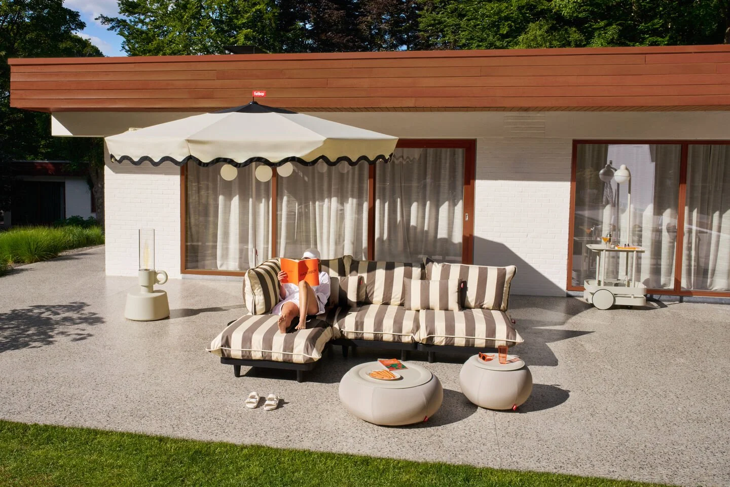 Person sitzt auf einer Strandliege im Garten, liest ein Buch, bei Sonnenschein, mit einem Regenschirm und stilvollem Outdoor-Mobiliar.