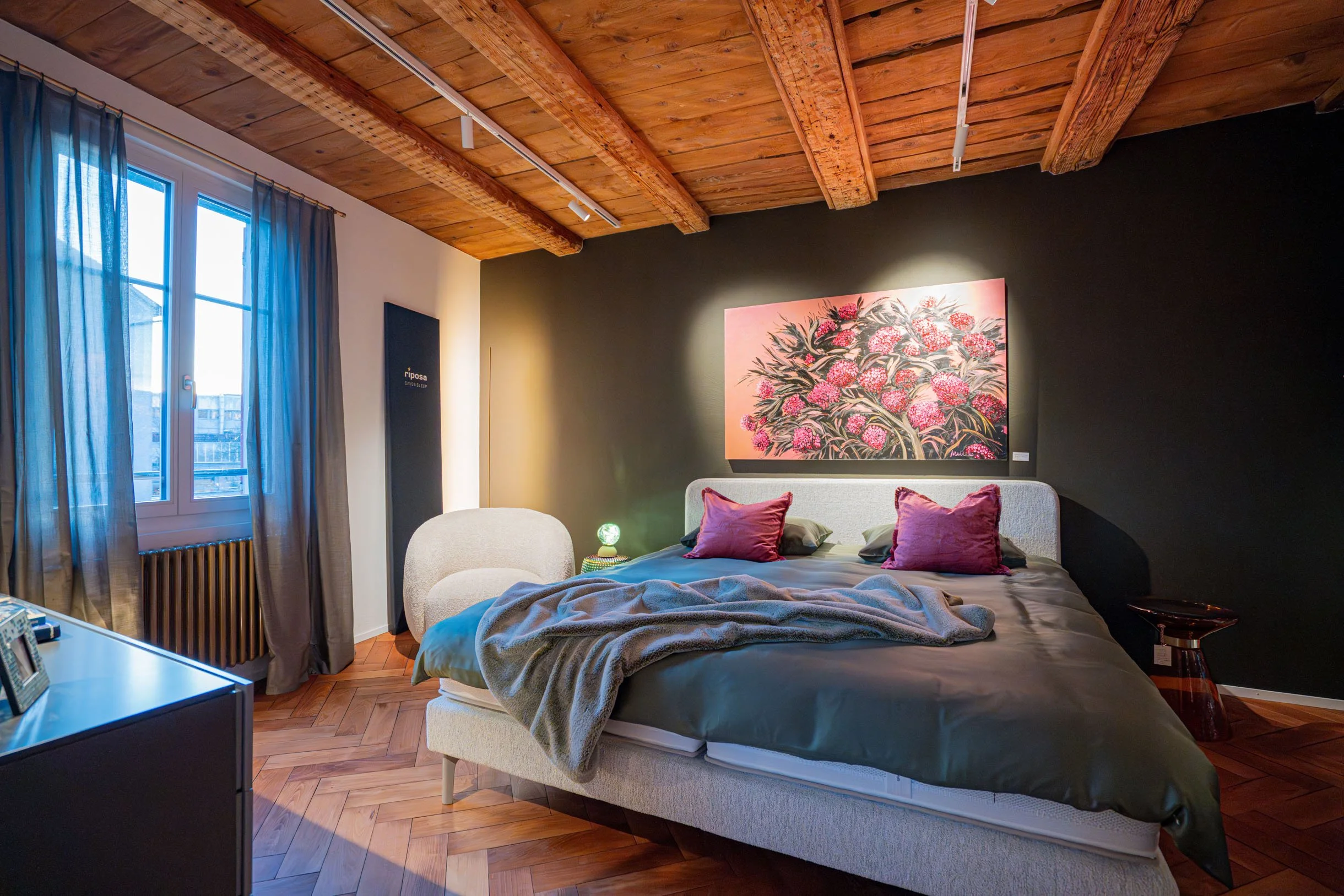 Ein Schlafzimmer mit Holzdecke, grauer Wand, Bett mit grauem Bezug und roten Kissen, Wandkunst mit Blumenmotiven, seitlicher Nachttisch, Fenster mit blauen Vorhängen.