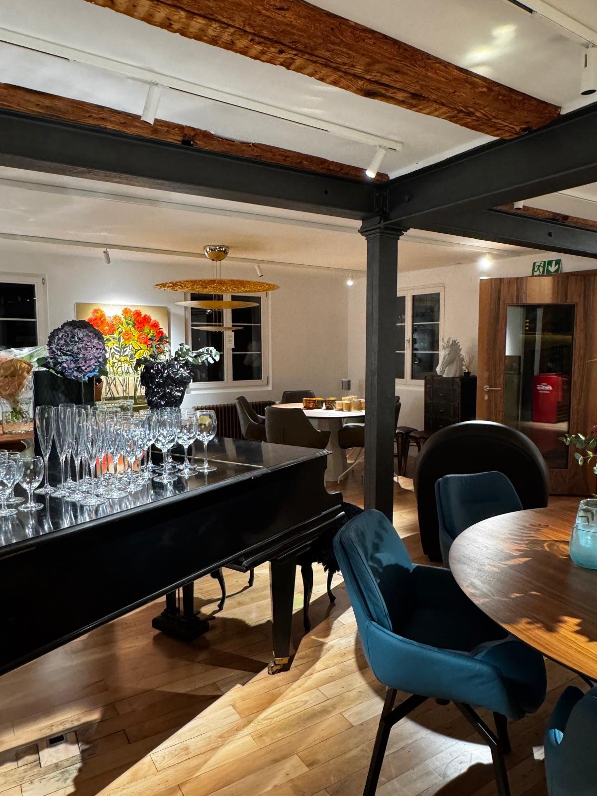 Ein gemütliches Esszimmer mit einem Klavier, auf dem zahlreiche Sektgläser stehen, einem runden Holztisch mit blauen und schwarzen Sesseln, einer schwarzen Kommode, einem Gemälde mit roten Blumen an der Wand, geöffneten Fenstern und Holzbalken an der