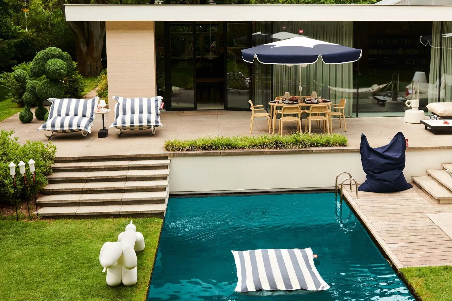 Ein moderner Außenbereich mit Swimmingpool, Liegen mit gestreiften Kissen, einem rundem Gartentisch mit Stühlen unter einem Sonnenschirm und einem Sessel unter einem großen Sonnenschirm. Es gibt auch einen kunstvollen weißen Dekorations-Ballon-Hund und eine skulpturartige Figur. Das Haus hat große Fenster und ist von grünen Büschen und Bäumen umgeben.