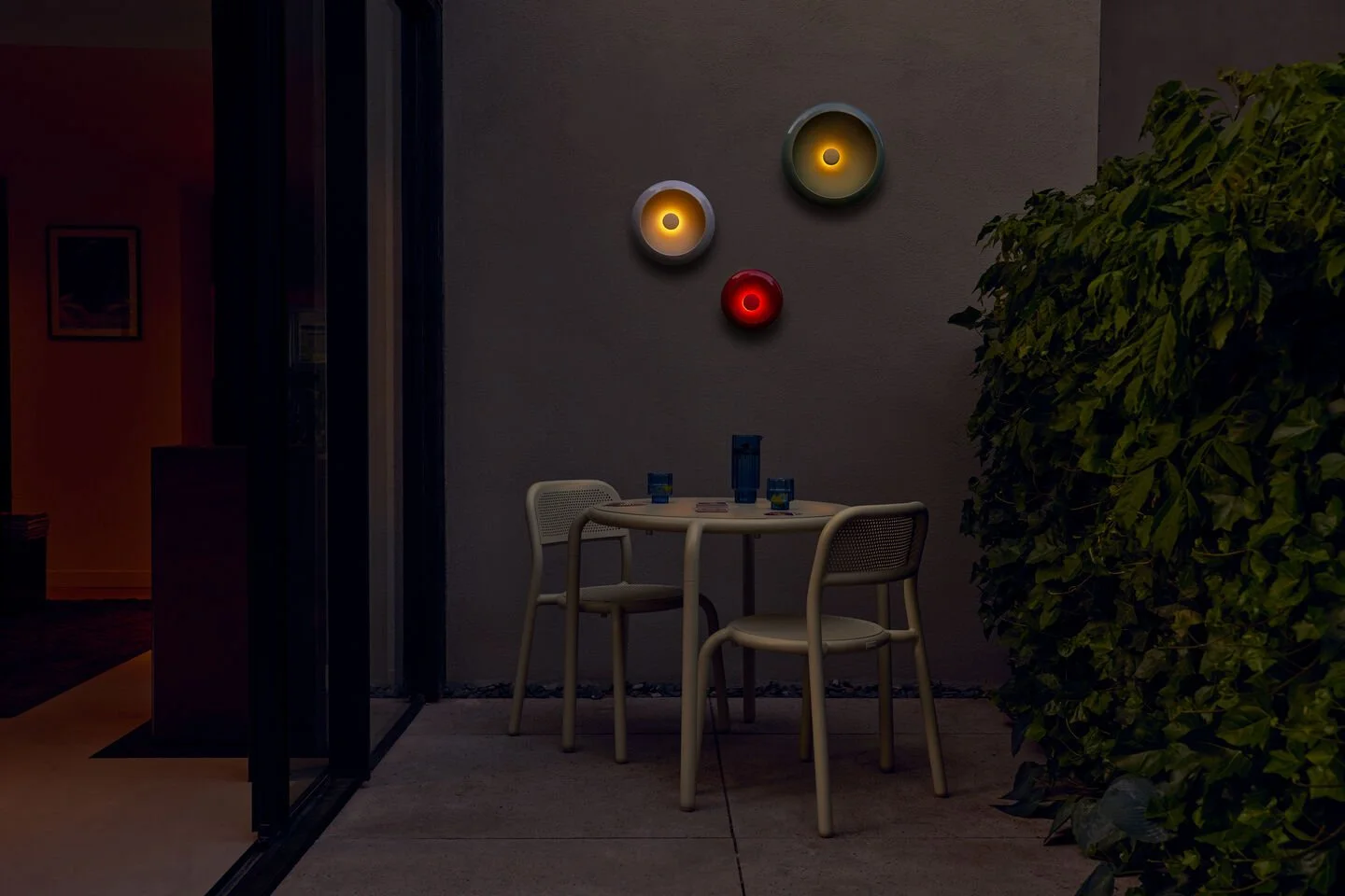 Ein dunkler Balkon mit einem runden Tisch, drei Stühlen und einer großen Zimmerpflanze auf der rechten Seite. An der Wand hängen drei runde, farbige Lichter in Orange, Weiß und Rot.