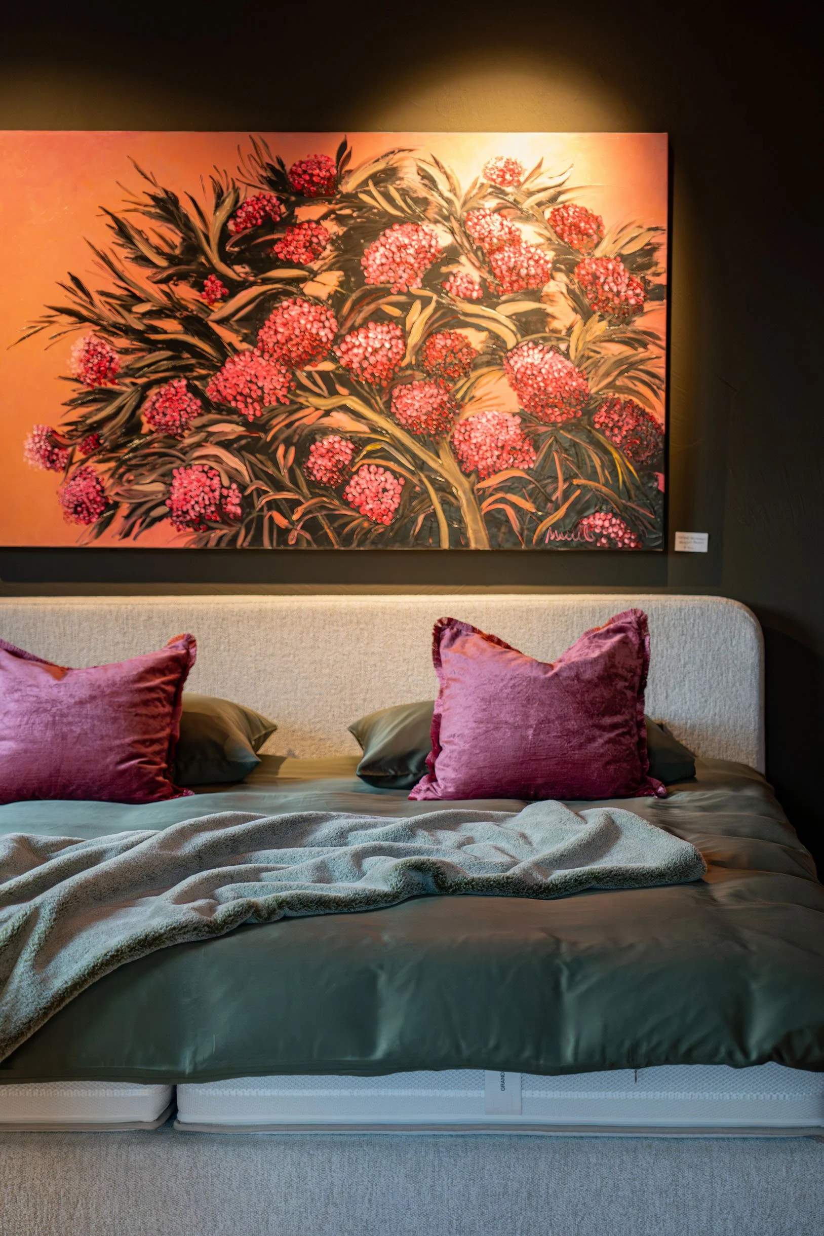 Ein Bett mit dunkelgrauem Bettzeug und mehreren Kissen in Rosa- und Grautönen, vor einem Wandbild mit pinken Blumen und grünen Blättern.