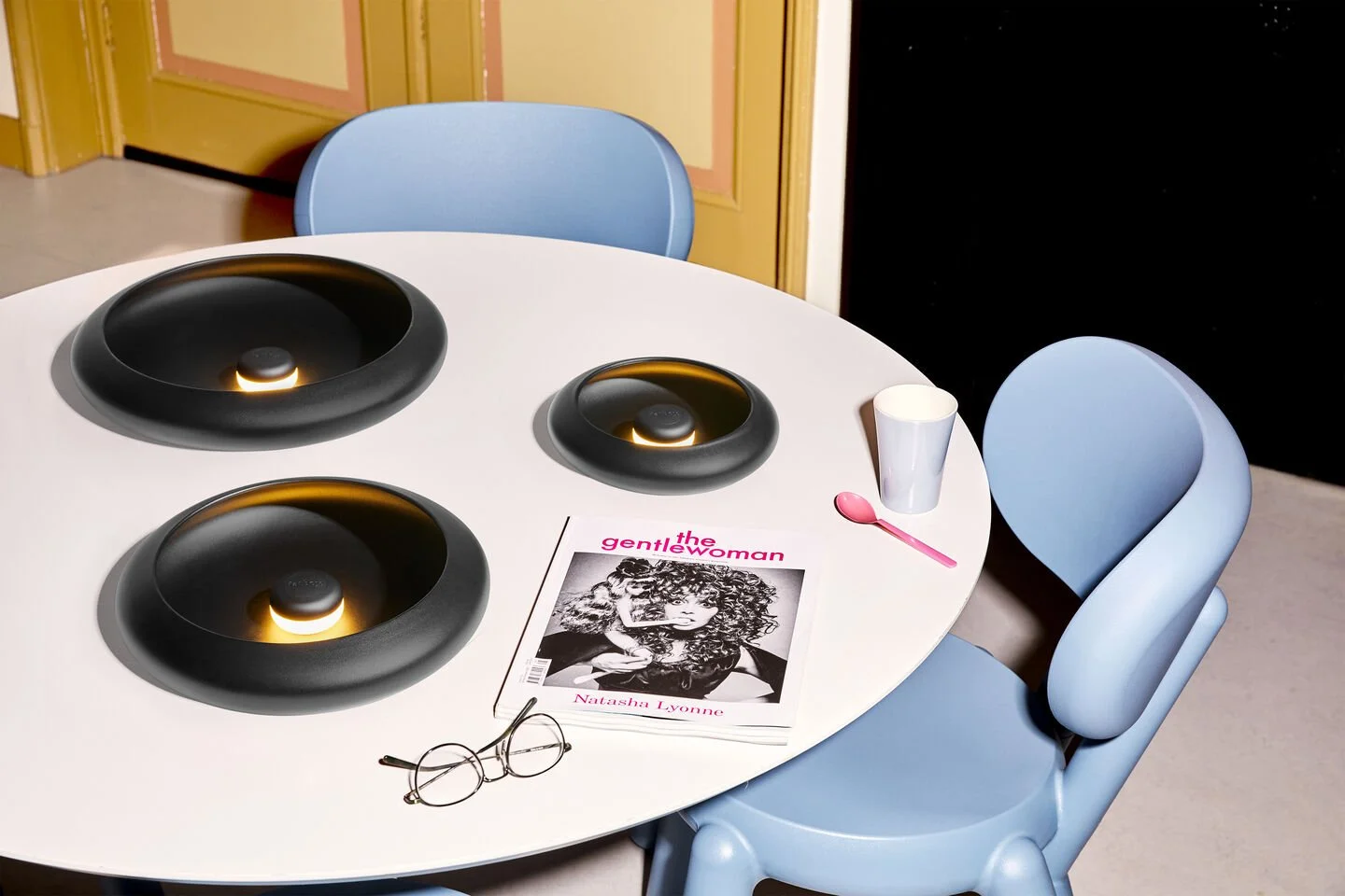 Ein runder Esstisch mit drei schwarzen Lautsprechern, einer weißen Tasse, einer rosa Löffel, einer Zeitschrift mit einem schwarzen und weißen Coverbild und einem Titel in pink, sowie einer Brille. Zwei blaue Stühle sind um den Tisch herum, im Hintergrund eine gelbe Tür.
