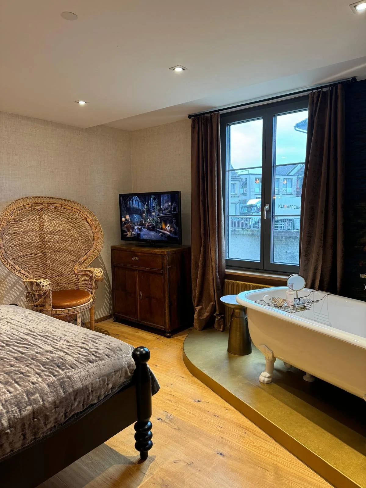 Ein Schlafzimmer mit einem schwarzen Bett, einem Holzschrank mit Fernseher, einem Korbsessel, einer Badewanne auf einem erhöhten goldfarbenen Podest und Fenster mit dunklen Vorhängen.