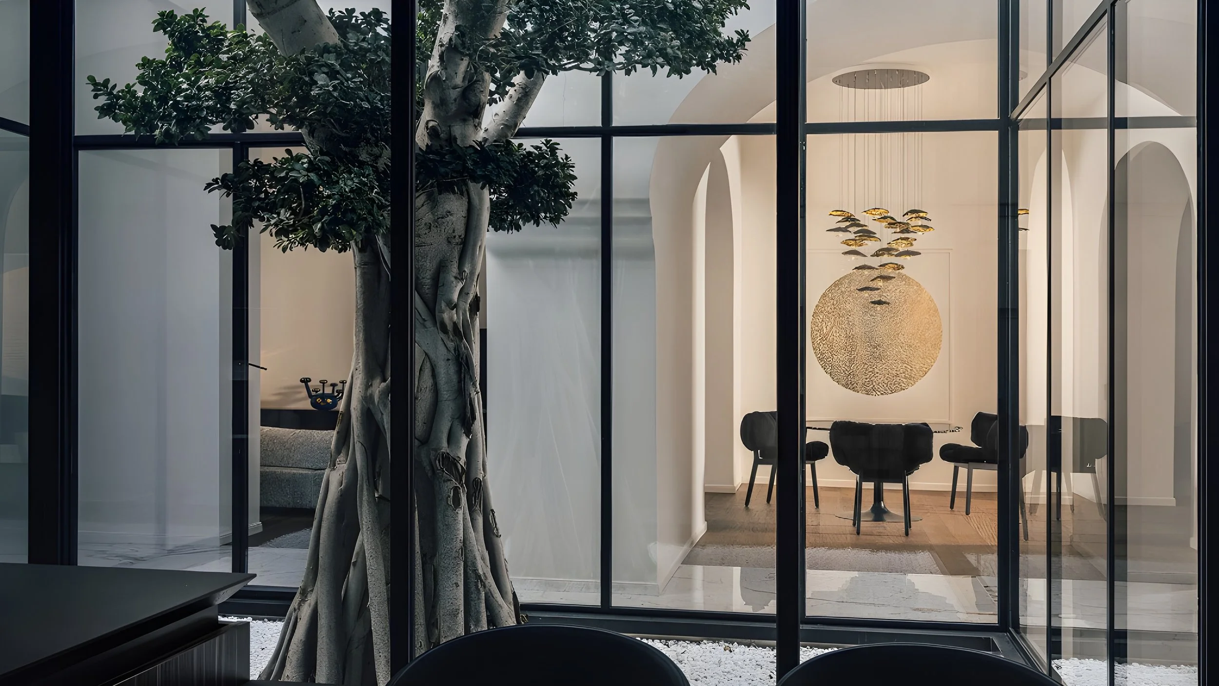 Innenraum eines modernen Hauses mit großen Glasfenstern, einem dekorativen Baum, einem Kronleuchter und einem Essbereich mit Stühlen.