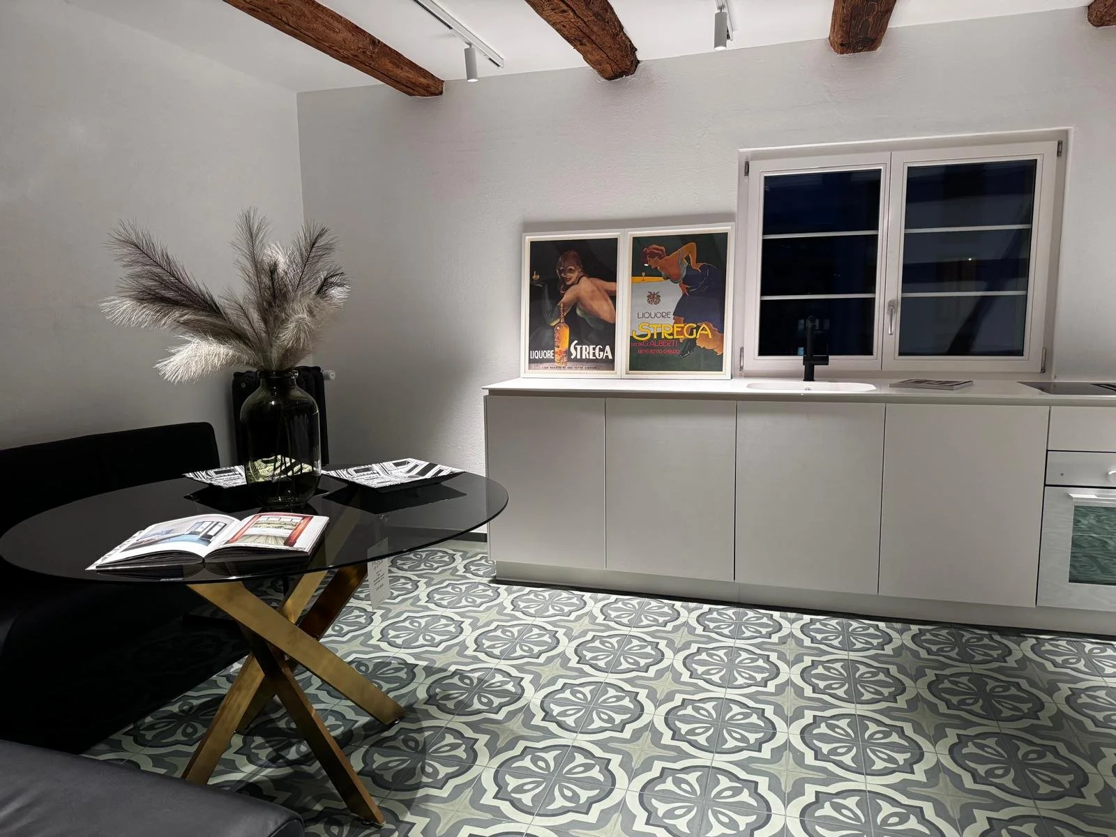 Modernes Wohnzimmer mit weißer Wand, dunklem Sofa, schwarzem Tisch mit geöffnetem Buch, Vase mit getrockneten Pflanzen, weißen Schränken, Fenster und dekorativen Postern an der Wand