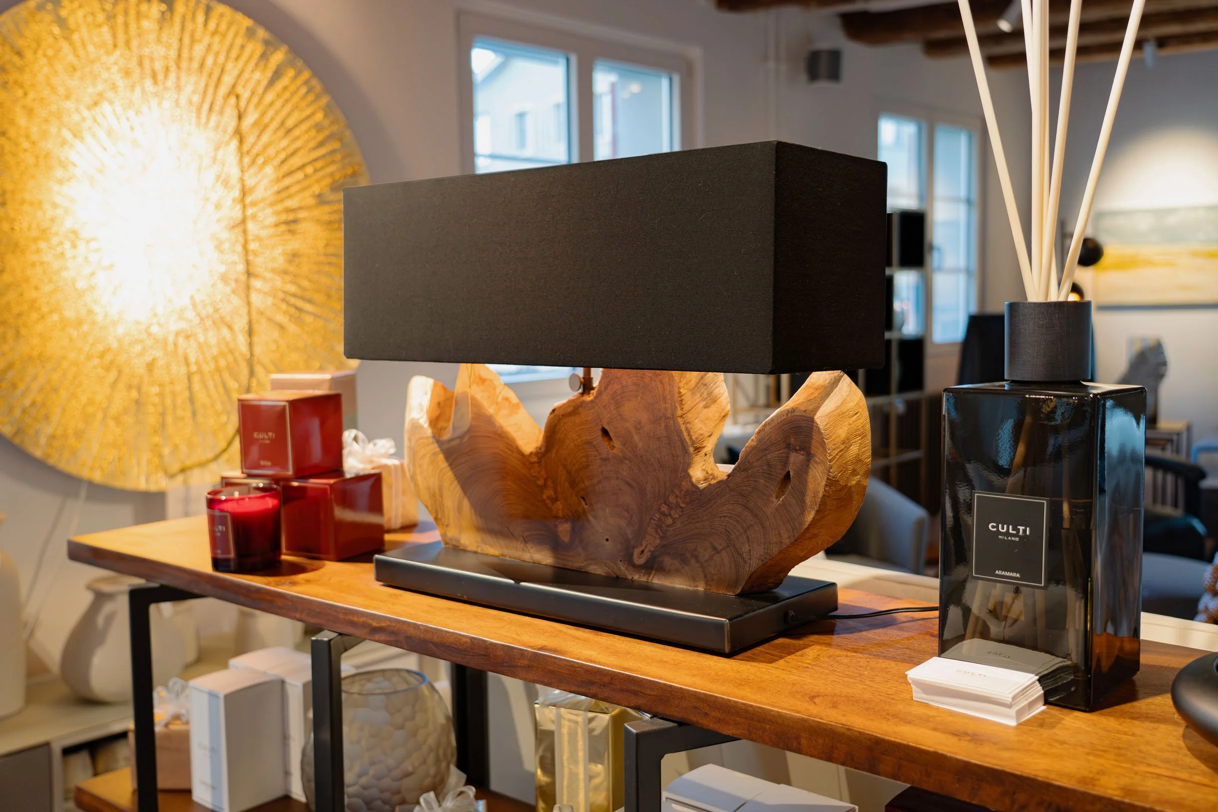 Ein Holztisch mit dekorativen Gegenständen, darunter eine schwarze Tischlampe auf einem Holzsockel, rote Kerzen, eine schwarze Vase mit weißen Zweigen und einige Broschüren. Im Hintergrund sind Fenster und weitere Inneneinrichtung zu sehen.