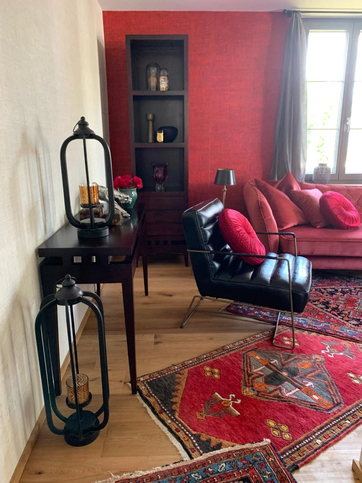 Ein Wohnzimmer mit rotem Wandteppich, schwarzem Sessel mit roter Kissen, einer roten Couch mit mehreren roten Kissen, einem schwarzen Tischlampe, einer Kommode mit Blumen, Dekorationen und einem Fenster mit grauen Gardinen.