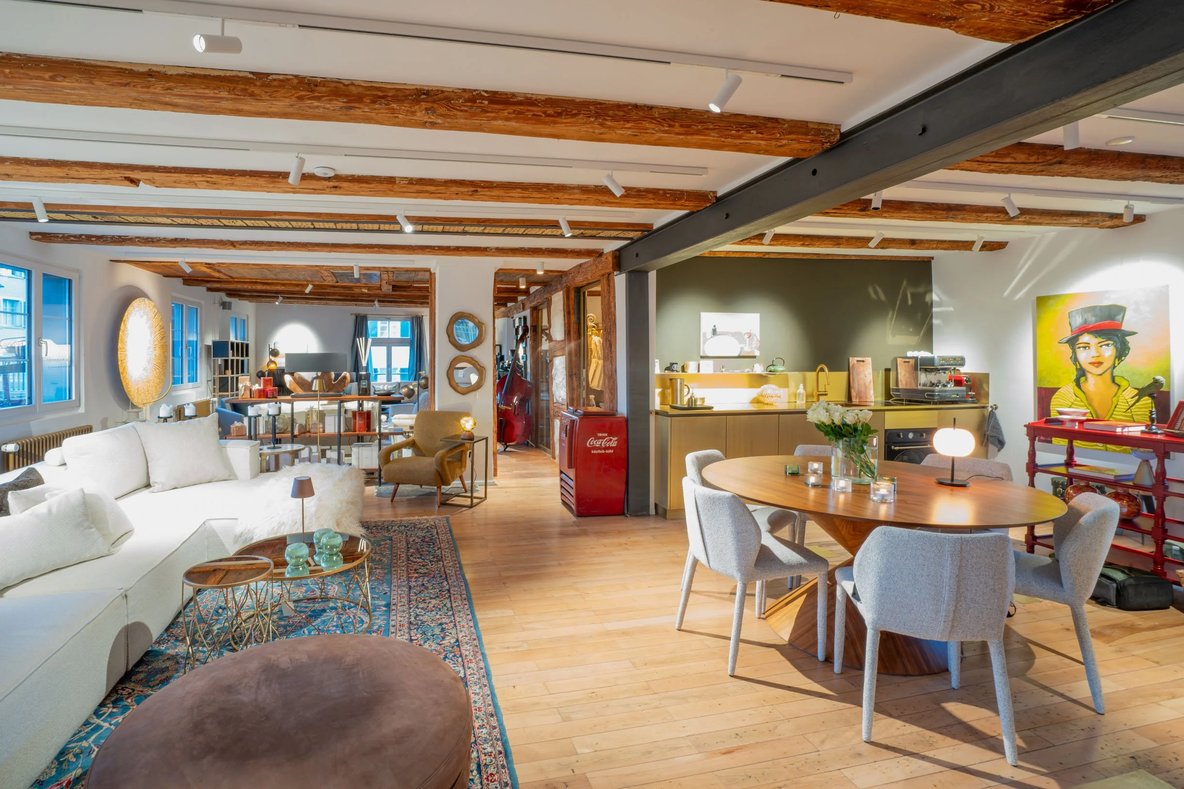 Ein modernes, offen gestaltetes Wohnzimmer mit Holzdeckenbalken, großen Fenstern, weißen Wänden, einem weißen Sofa, mehreren Sesseln, einem Esstisch mit Stühlen, Kunstwerken an den Wänden und verschiedenen Wohn- und Dekorationsartikeln.