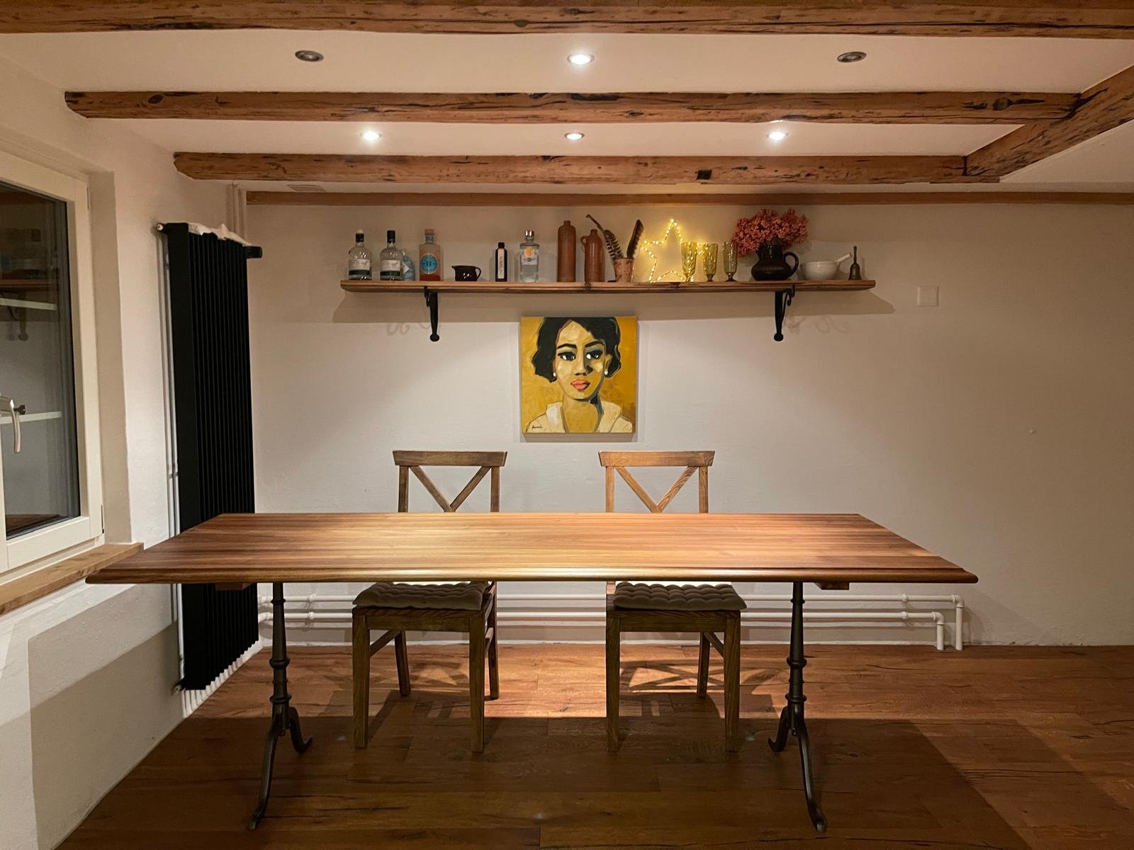 Ein Esszimmertisch mit zwei Stühlen vor einer Wand, die ein Portrait einer Frau und eine schwarze Wandlampe zeigt. Oben eine Holzregal mit dekorativen Gegenständen und Lichterstern.