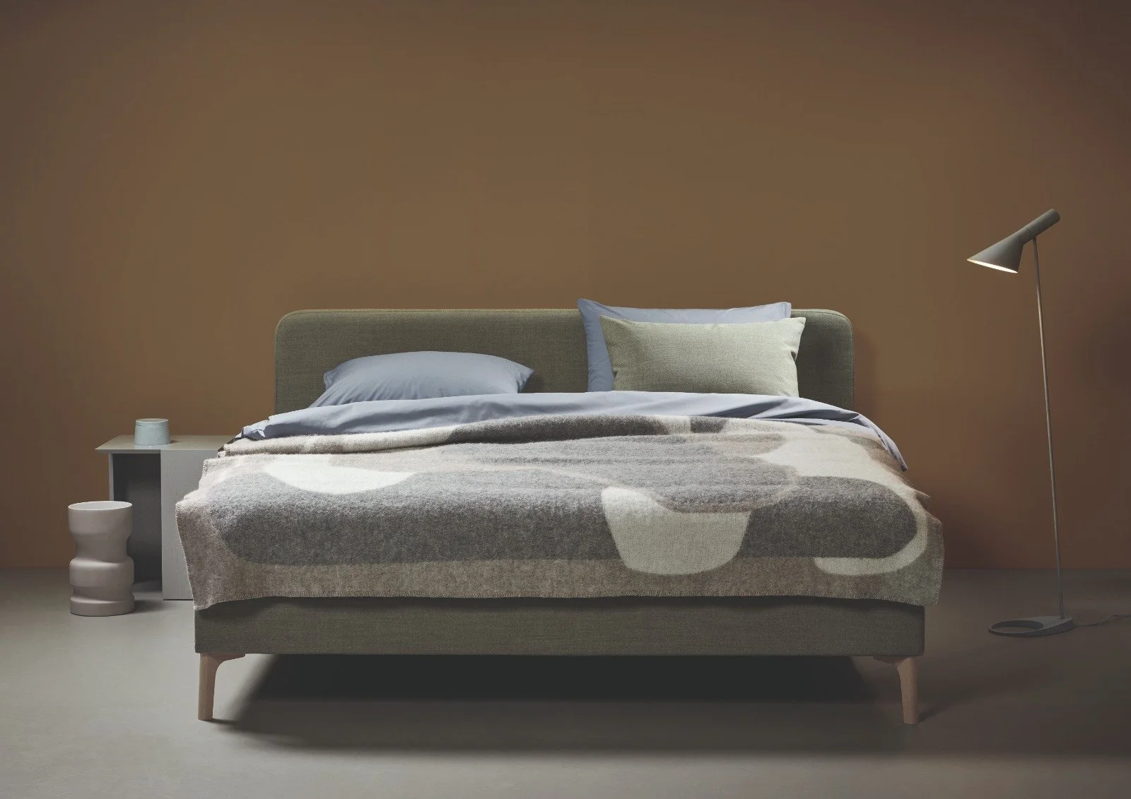 Ein modernes Schlafzimmer mit einem grauen Bett, das mit einer gemusterten Decke bedeckt ist. Es gibt zwei Kissen, eines in Grau und eines in Beige. Neben dem Bett steht ein kleiner Nachttisch mit einem weißen Becher, und auf der rechten Seite befindet sich eine Stehlampe mit einem nach unten gerichteten Licht. Die Wand im Hintergrund ist braun.