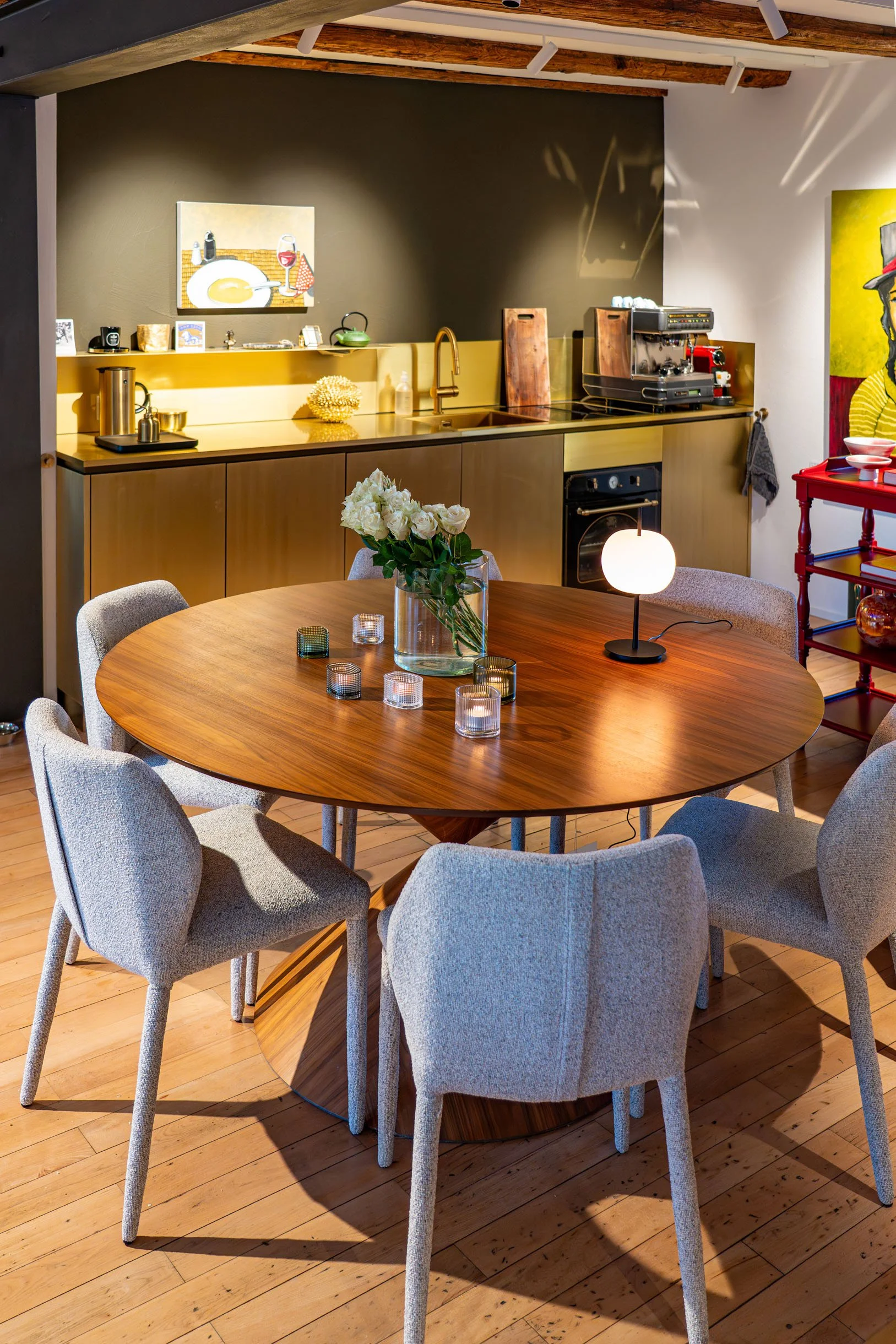 Ein runder Holz Esstisch mit sechs hellgrauen gepolsterten Stühlen, eine Vase mit weißen Rosen, mehrere kleine Kerzen, auf einem Holzboden. Im Hintergrund eine Küchenzeile mit goldfarbenen Schränken, einer Kaffeemaschine und dekorativen Gegenständen,