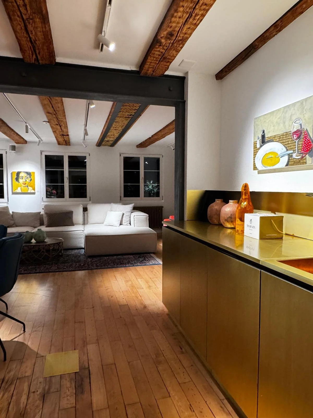 Modernes Wohnzimmer mit weißem Sofa, Holzdeckenbalken, Wanddekorationen und einem goldenen Küchenregal mit Vasen und Dekorationsstücken.