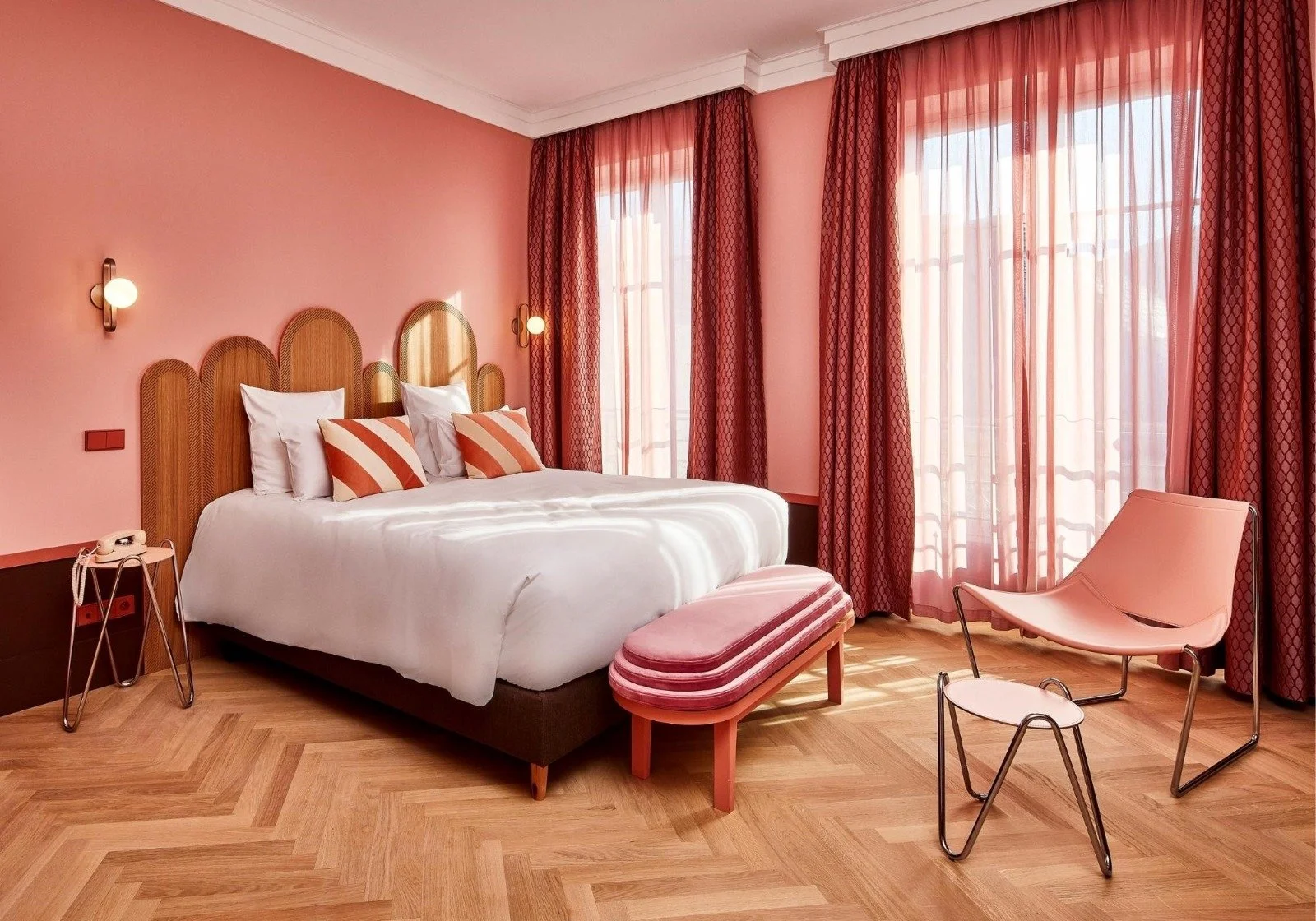 Ein Hotelzimmer mit pinkfarbenen Wänden, großen Fenstern mit durchsichtigen und roten Vorhängen, einem weiß bezogenen Bett mit mehreren weißen und orange-gestreiften Kissen, ein pinkfarbener Stuhl und ein kleiner Tisch, elegante Beleuchtung.