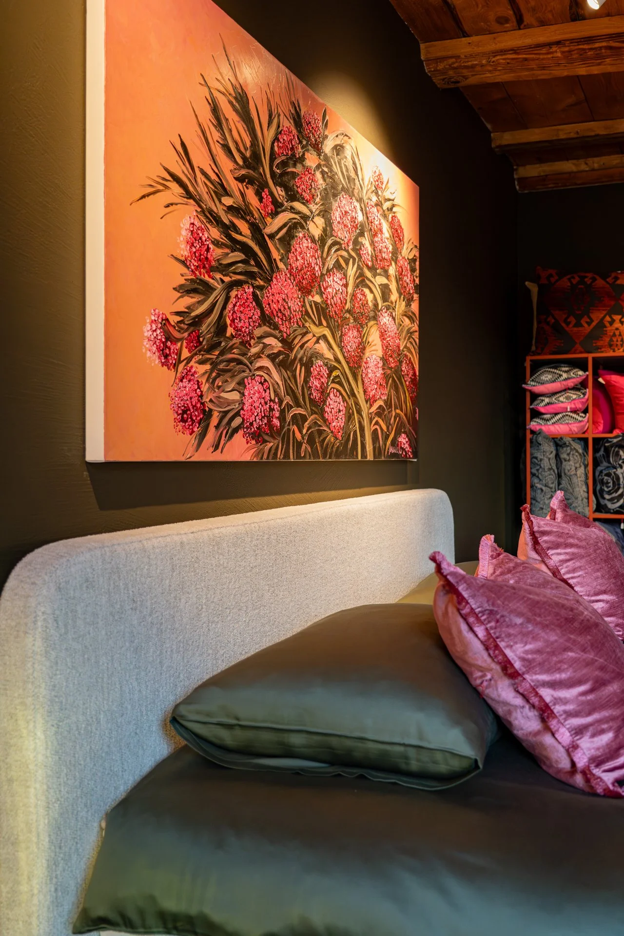 Ein Wohnzimmer mit einem hellgrauen Sofa, pinken Kissen, einem schwarzen Kissen und einer Wandkunst mit Blumenmustern. Die Wand ist dunkel, das Deckenholz ist sichtbar.