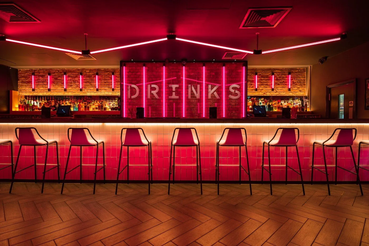 Innenaufnahme einer Bar mit Neonbeleuchtung in Pink und Rot, Barhockern, einem Brickwänden und der Aufschrift 'DRINKS' auf einer Wand mit Neonlinien.