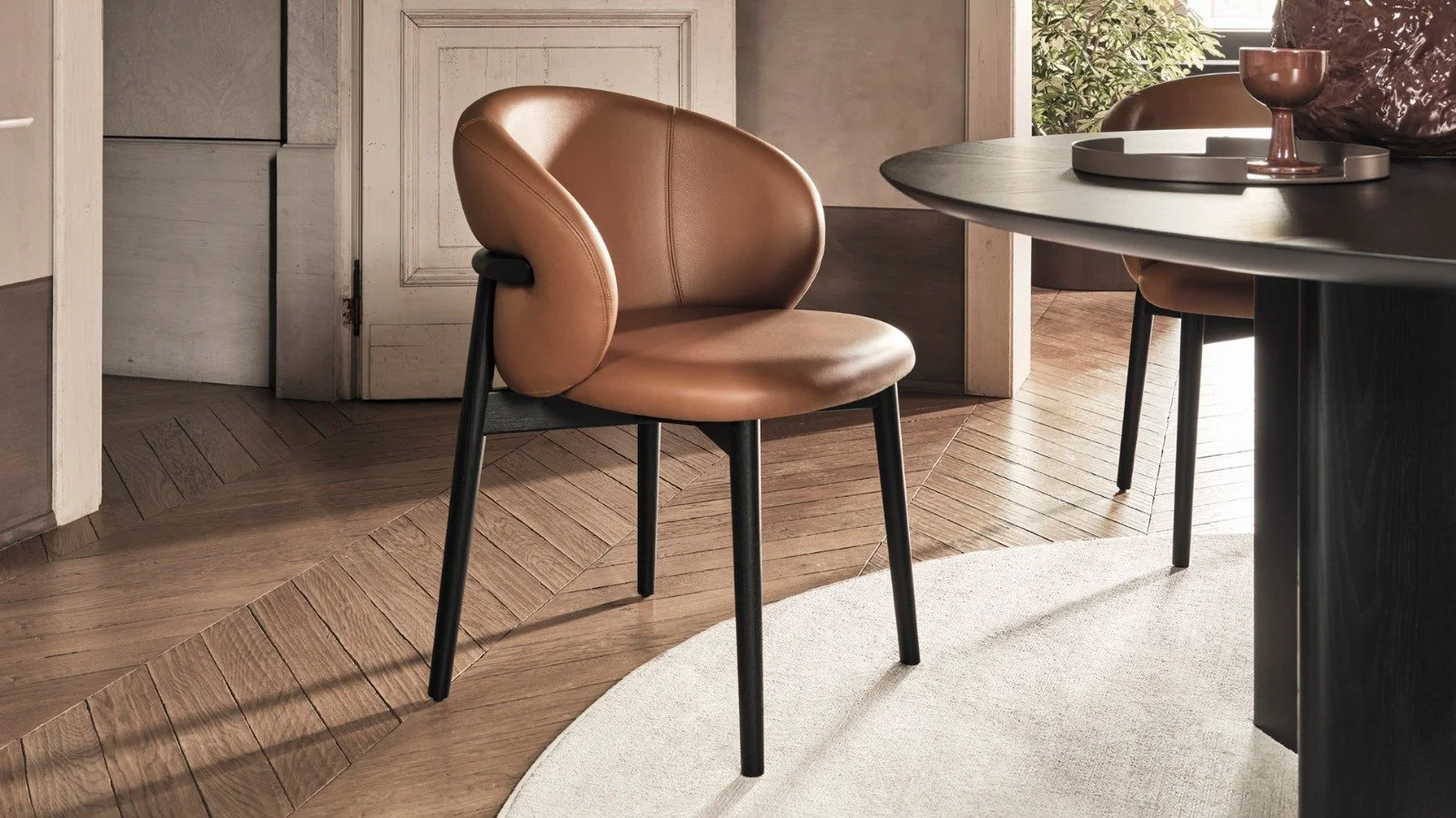 Ein moderner Sessel mit braunem Leder und schwarzen Beinen, im Esszimmer neben einem schwarzen Tisch mit Dekoration und einer Vase auf einem runden Teppich.