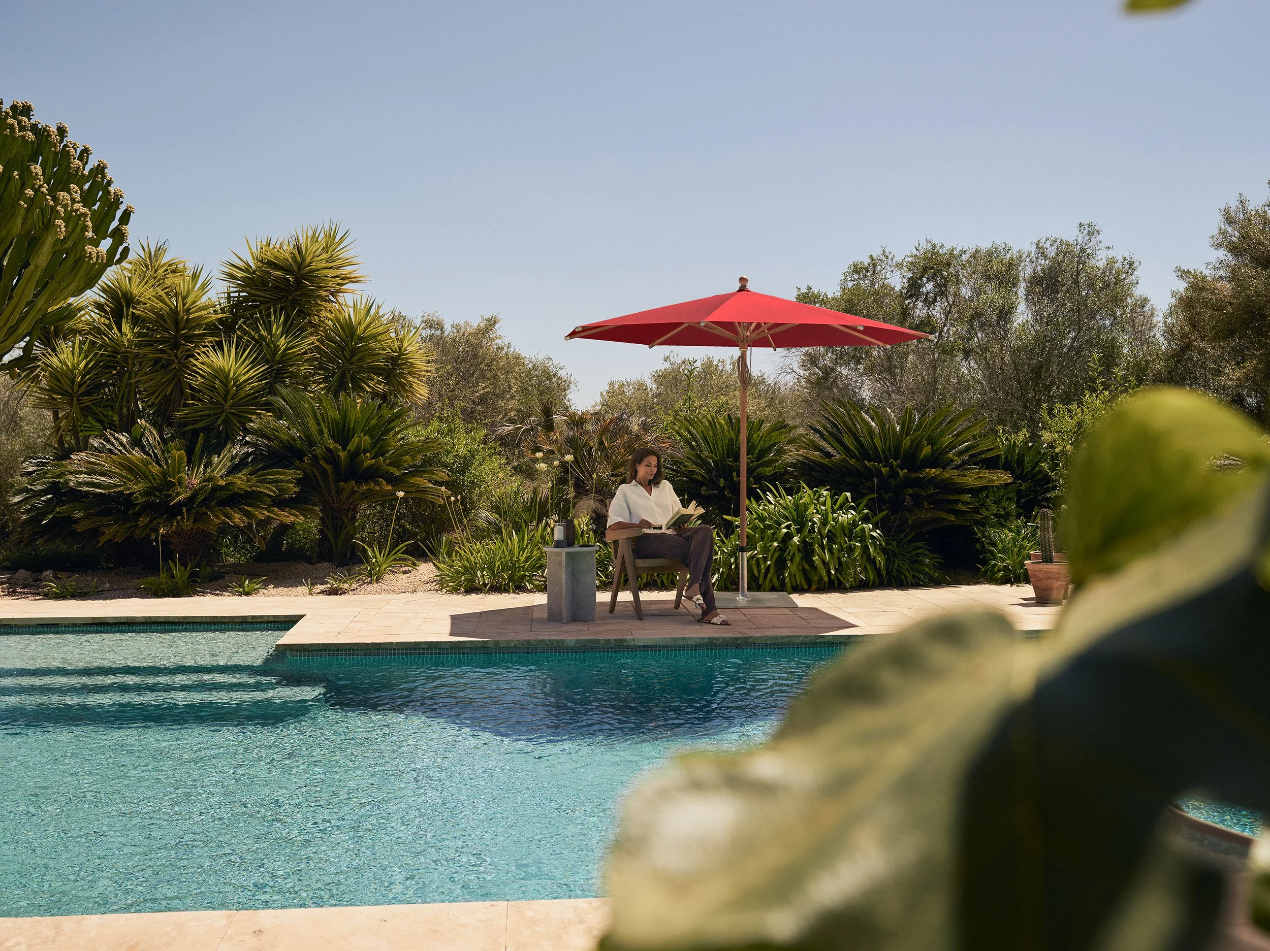 Frau sitzt unter einem roten Sonnenschirm am Pool und liest ein Buch, umgeben von grünen Pflanzen und Bäumen.