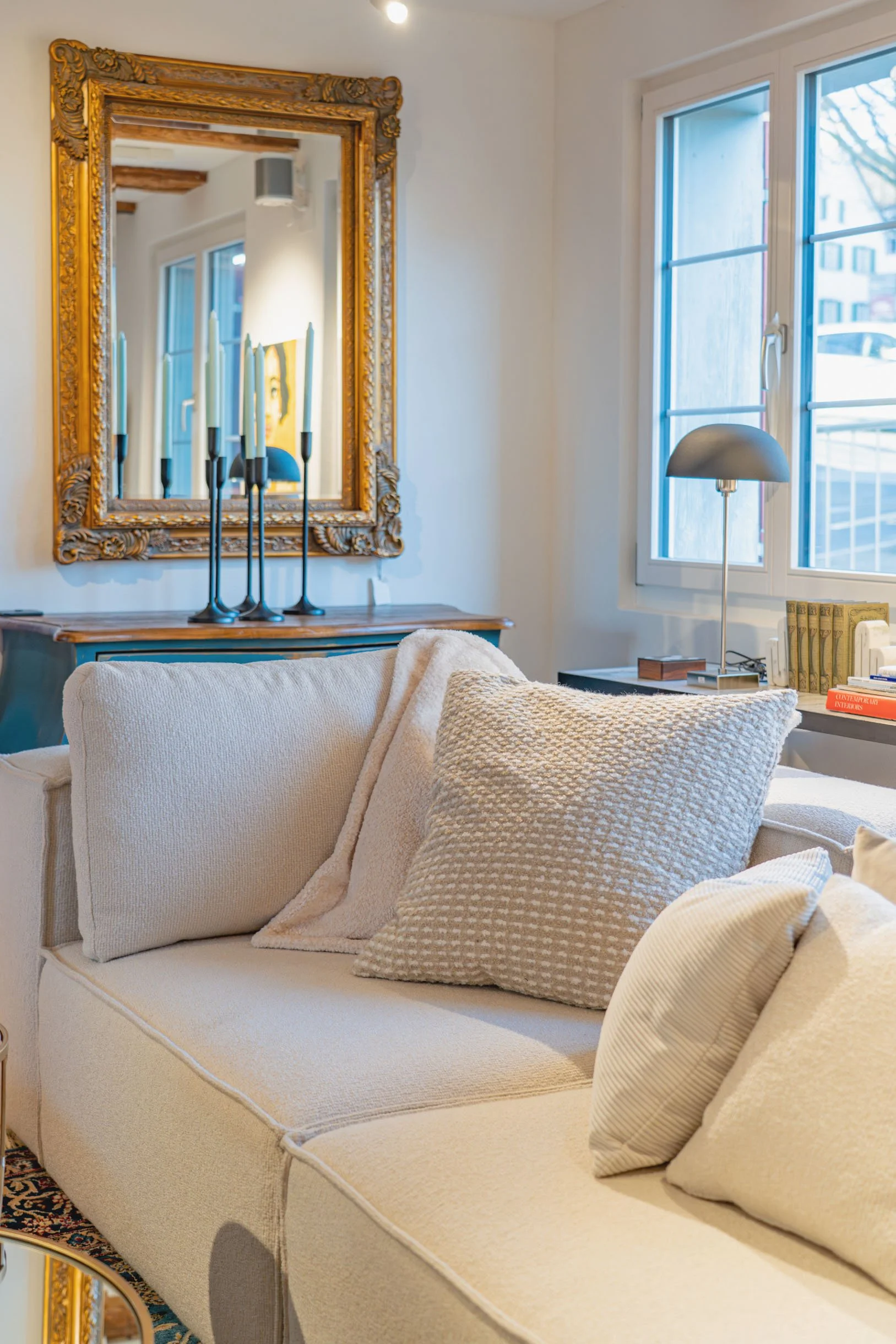 Ein Wohnzimmer mit einem weißen Sofa, mehreren Kissen, einem Fenster, einem Spiegel mit goldener Verzierung und einer Stehlampe.