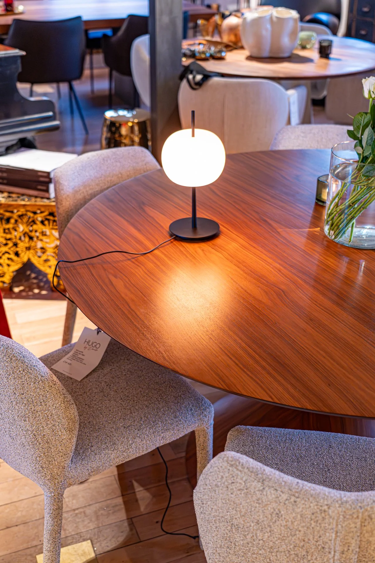 Ein runder Holztisch mit einer weißen Tischlampe, umgeben von beige, gepolsterten Esszimmerstühlen, in einem stilvollen, gemütlichen Raum.