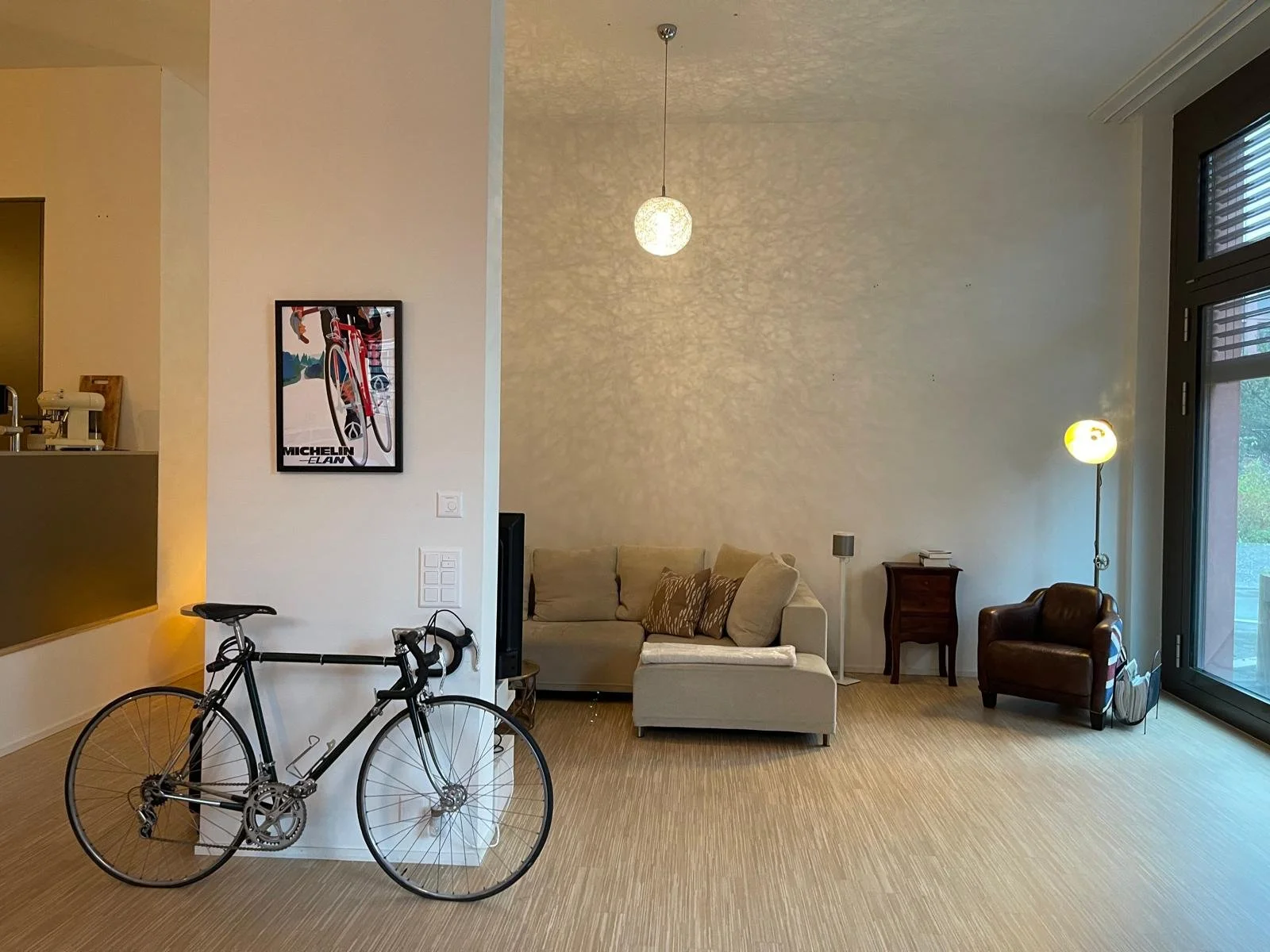 Ein Wohnzimmer mit Sofa, Sessel, Stehlampe, Wandbild, Fahrrad im Vordergrund und einer großen Fenstertür auf der rechten Seite