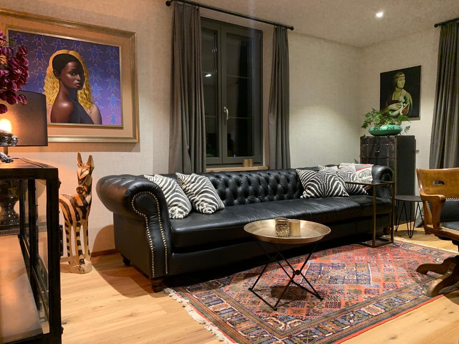 Innenaufnahme eines Wohnzimmers mit schwarzem Ledersofa, Zierkissen mit Tiermustern, buntem Teppich, Kunstwerken an den Wänden, großen Fenstern mit grauen Vorhängen, und verschiedenen Dekorationsgegenständen wie Zebrafigur, Lampen und Pflanzen.