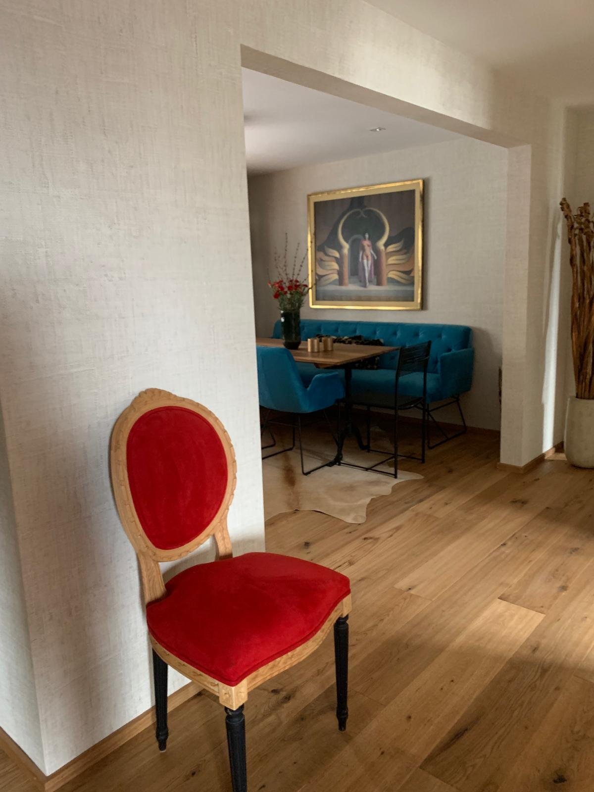 Ein roter Polsterstuhl im Vordergrund, im Hintergrund ein Essbereich mit blauen Sitzbänken, einem Holztisch, einem Blumenstrauß und einer großen Wandkunst mit goldfarbenem Rahmen.