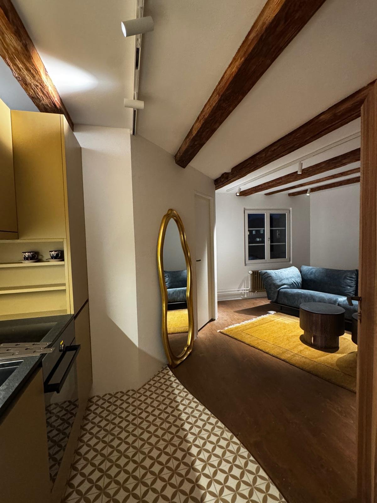 Ein Wohnzimmer mit Holzdecken, einem dunkelblauen Sofa, einem goldfarbenen Spiegel, einem gelben Teppich und einem kleinen schwarzen Tisch. Ein Fenster ist sichtbar.