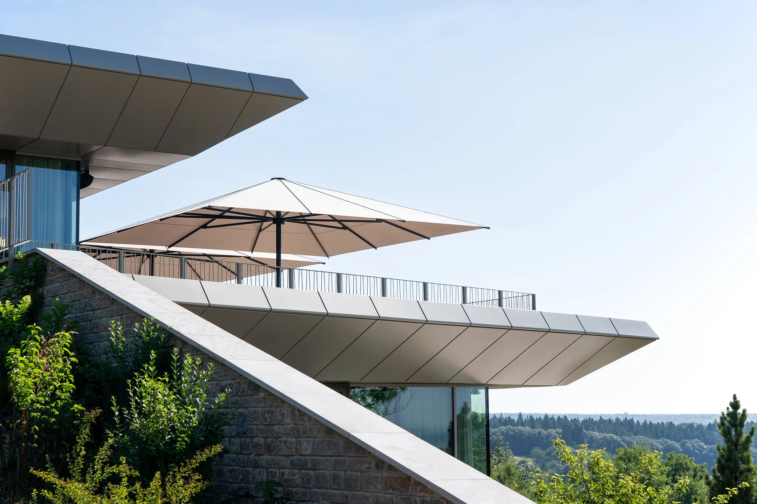 Moderne Gebäude mit großen Fensterflächen, schwarzen Metallgeländern, hellen Sonnenschirmen auf einer Terrasse, umgeben von grüner Vegetation, bei klarem Himmel.