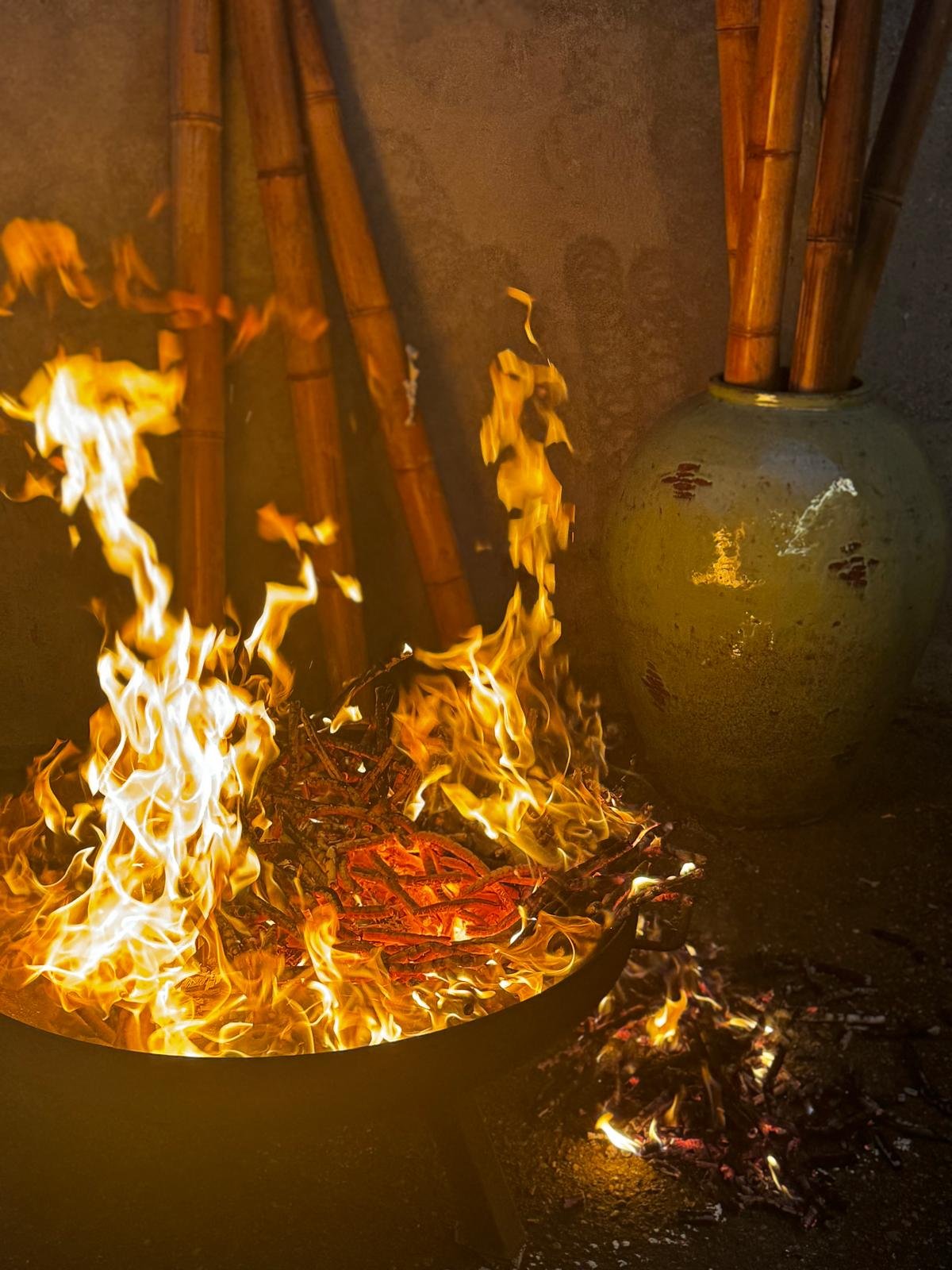Ein Lagerfeuer mit Flammen, das in einem Feuerkorb brennt, neben einem großen Keramikgefäß mit Holzstämmen an der Wand im Hintergrund.