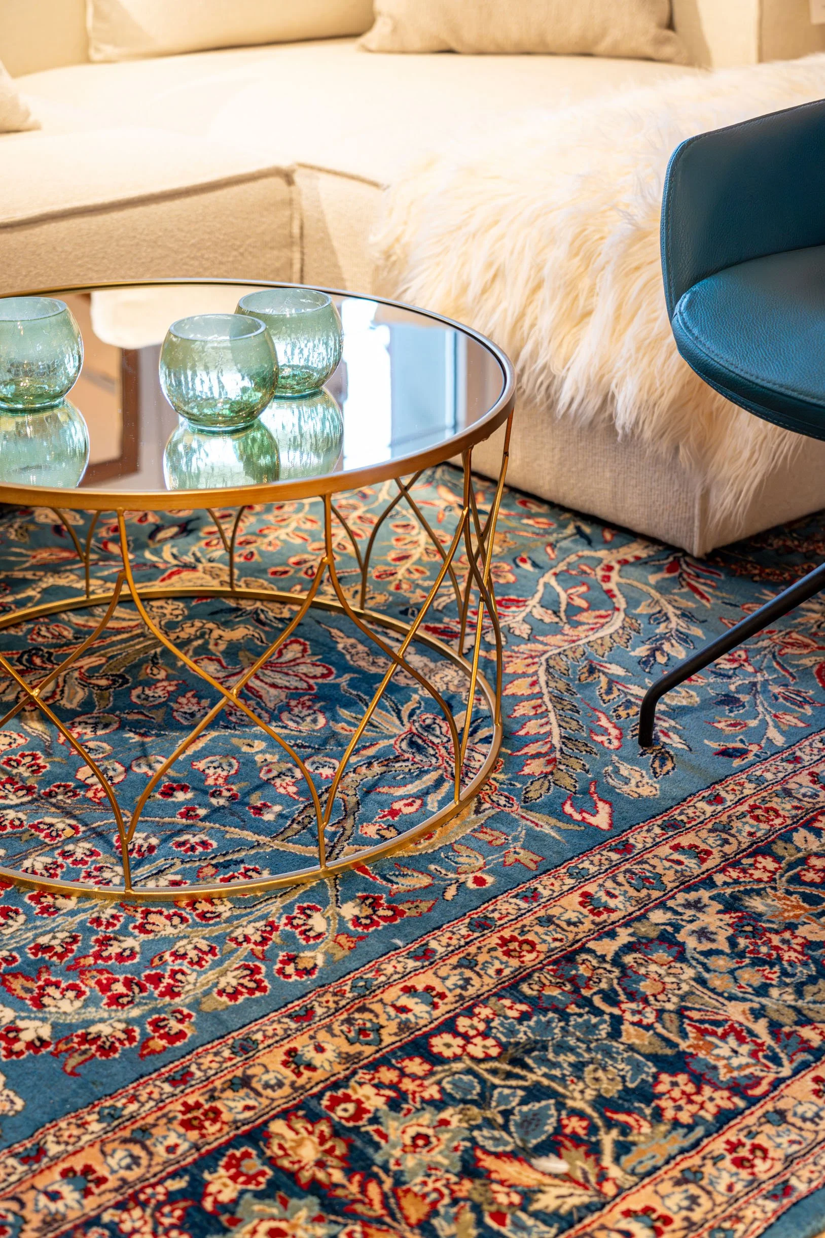 Ein Wohnzimmer mit einem blauen Sessel, einem runden Beistelltisch mit drei grünen Gläsern, einem Teppich mit floralem Muster und einem beige Couch mit Kissen.