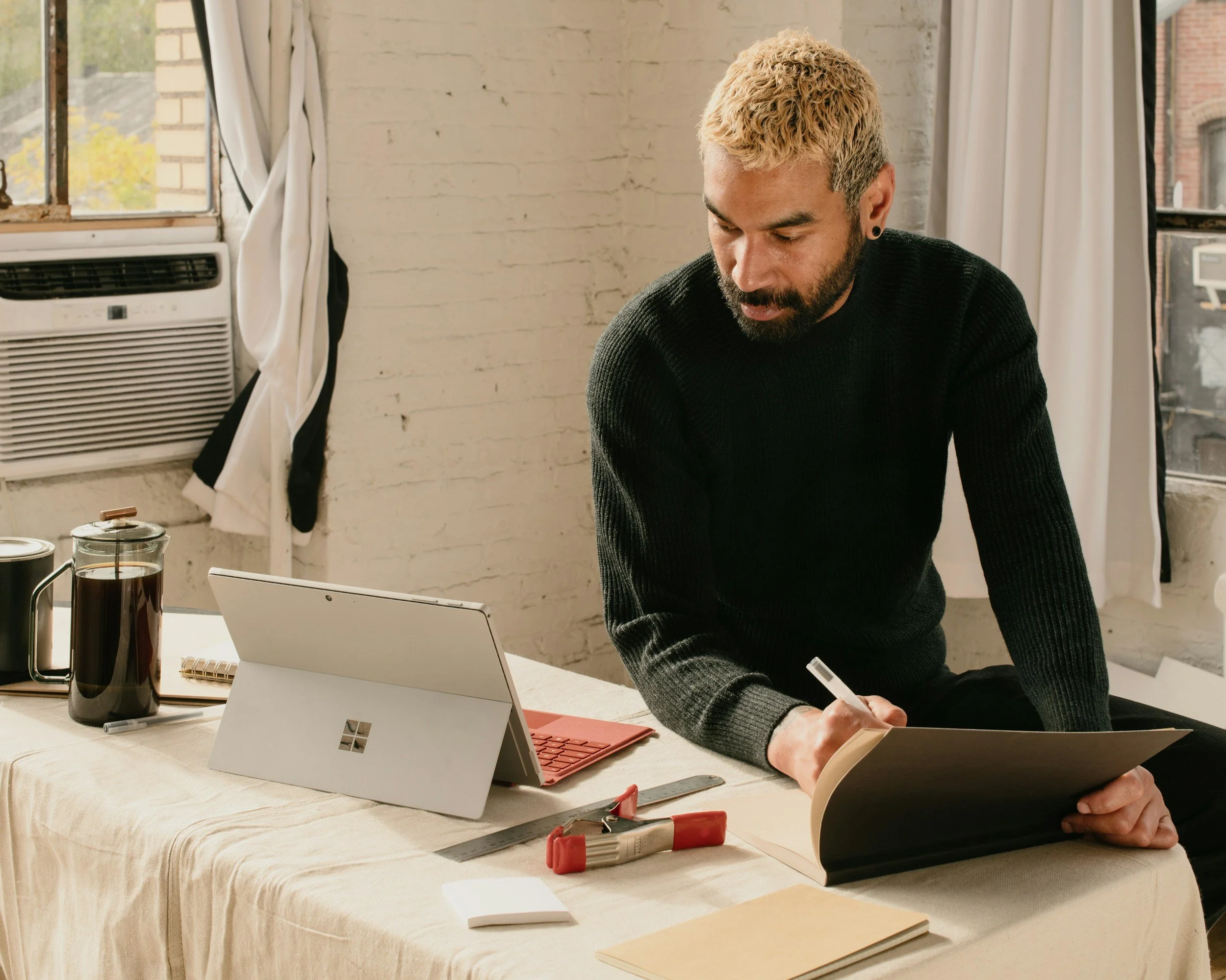 A man with light skin, blonde curly hair, and a beard, wearing a black sweater, is sitting at a table looking at a folder and writing on it with a marker. The table has a tablet, a mug, a pitcher, a notebook, a ruler, a stapler, and papers. There are windows with white curtains and an air conditioning unit in the background.