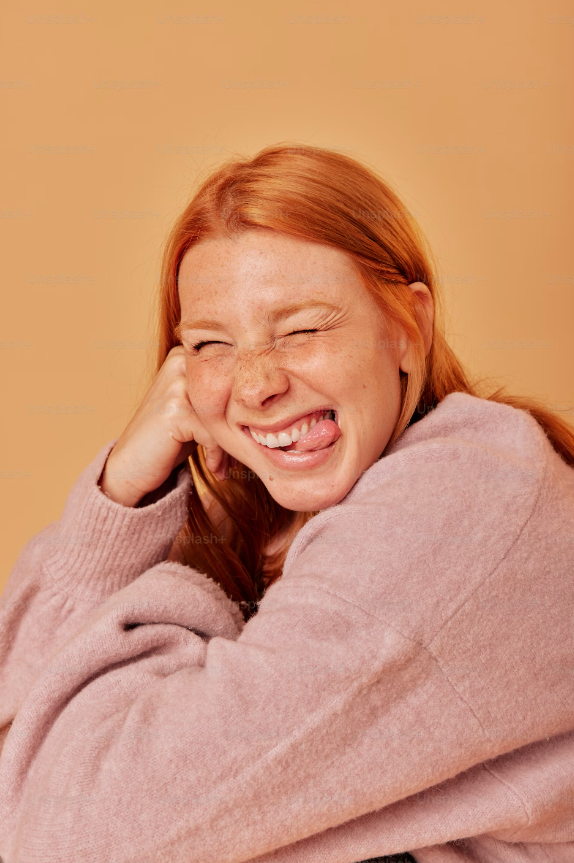Red-haired woman smiling with eyes closed and sticking out tongue, wearing a pink sweater, against a beige background.