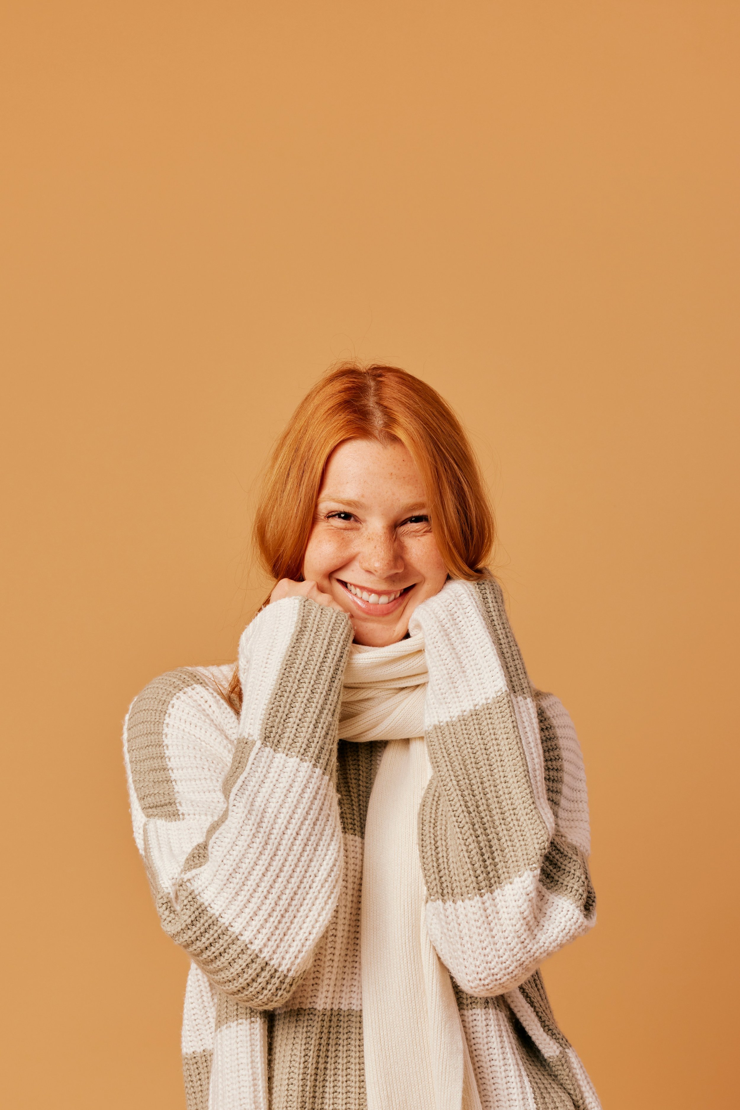 A young woman with red hair, freckles, and a bright smile, wearing a beige and white striped sweater and a cream scarf, standing against a warm beige background.