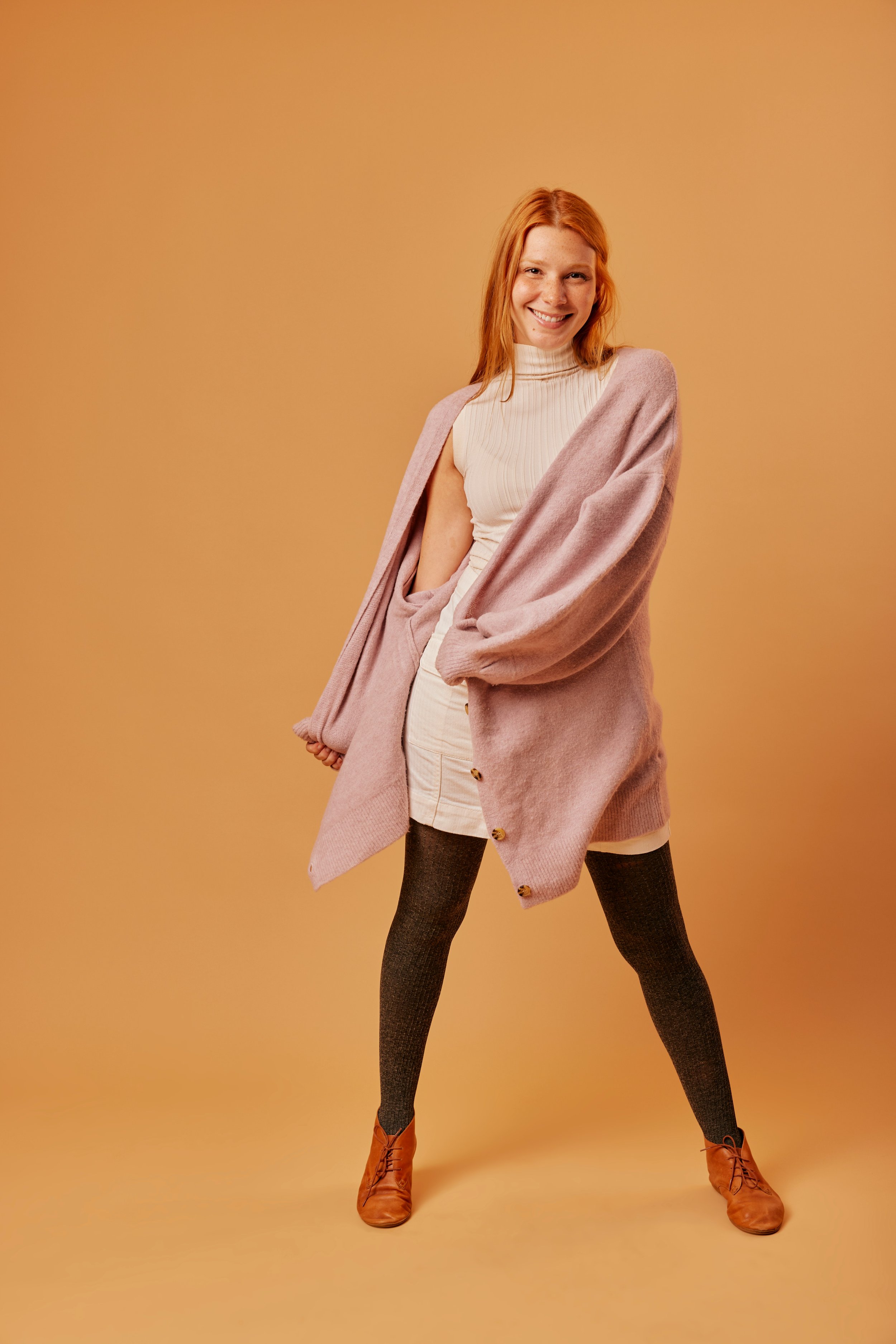 A woman with red hair wearing a beige turtleneck, a light pink oversized cardigan, a beige skirt, dark tights, and brown ankle boots, standing against a solid beige background, smiling confidently.