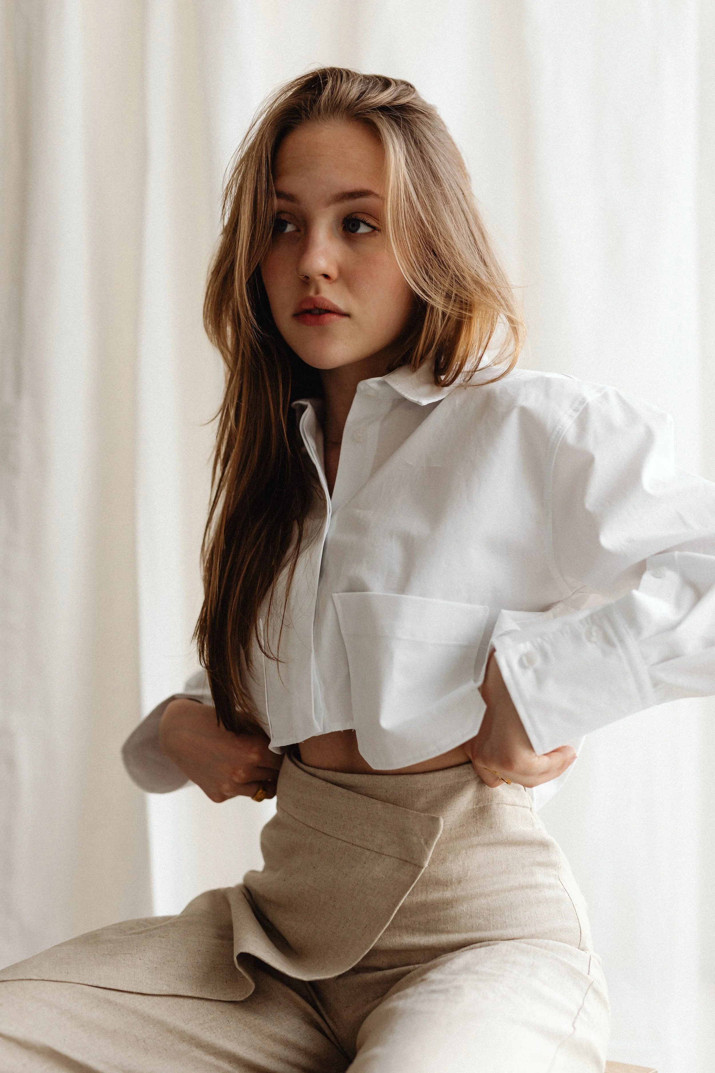A young woman with long, wavy brown hair wearing a white button-up shirt and beige pants, standing indoors near a light-colored curtain.