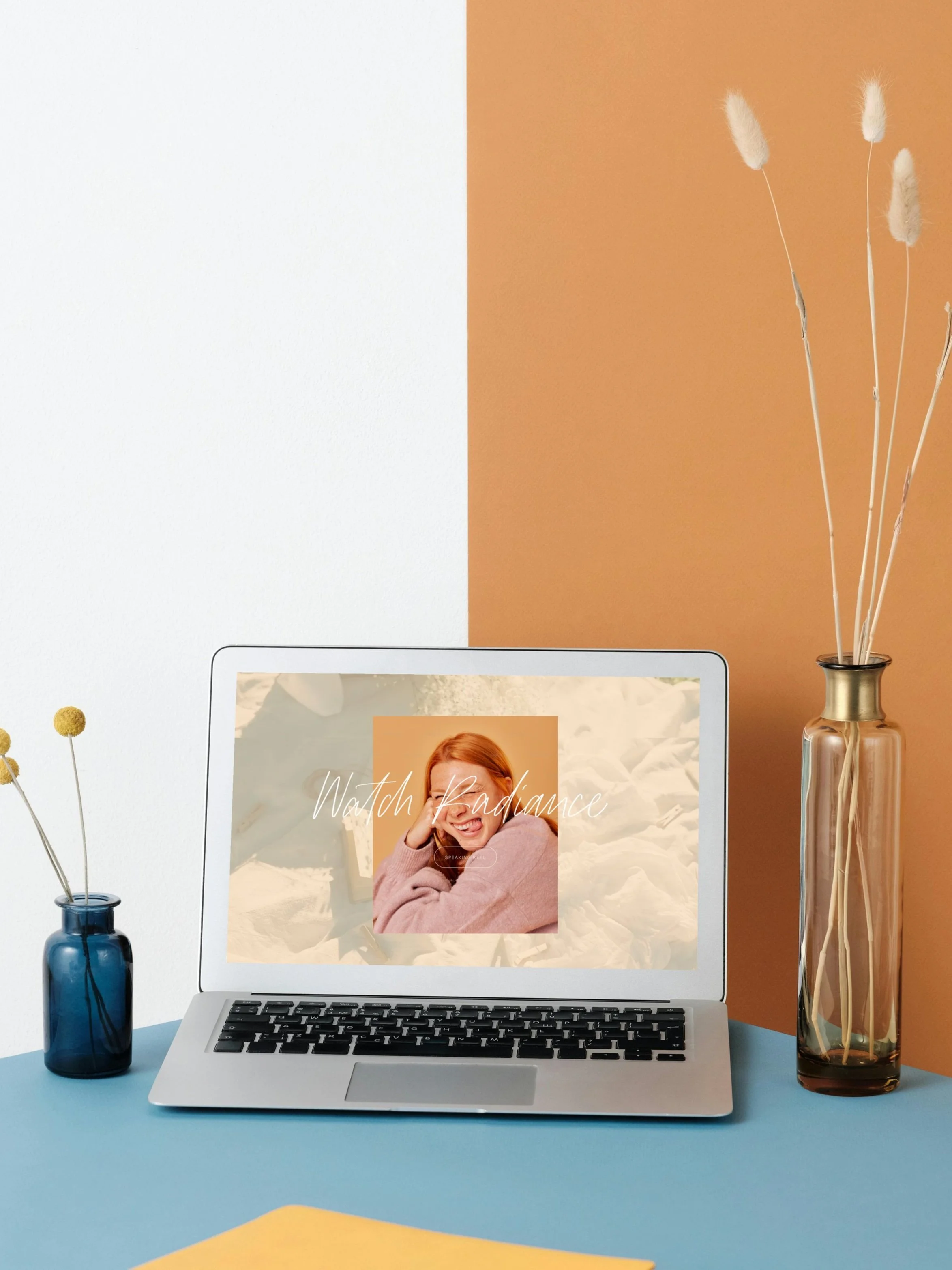 Open laptop on a blue desk with a screen that says 'Watch Radiance' and displays a smiling woman with red hair. To the left of the laptop, a small blue vase with yellow billy ball flowers. To the right, a tall brown vase with dried pampas grass. The background has a white wall on the left and a tan-colored wall on the right.