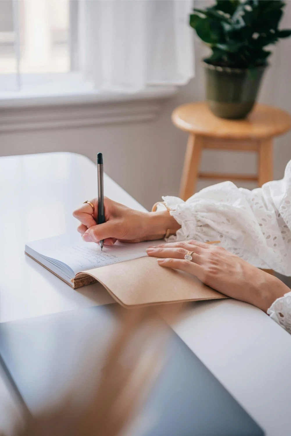 Person writing in a notebook at a white desk with a window and potted plant in the background