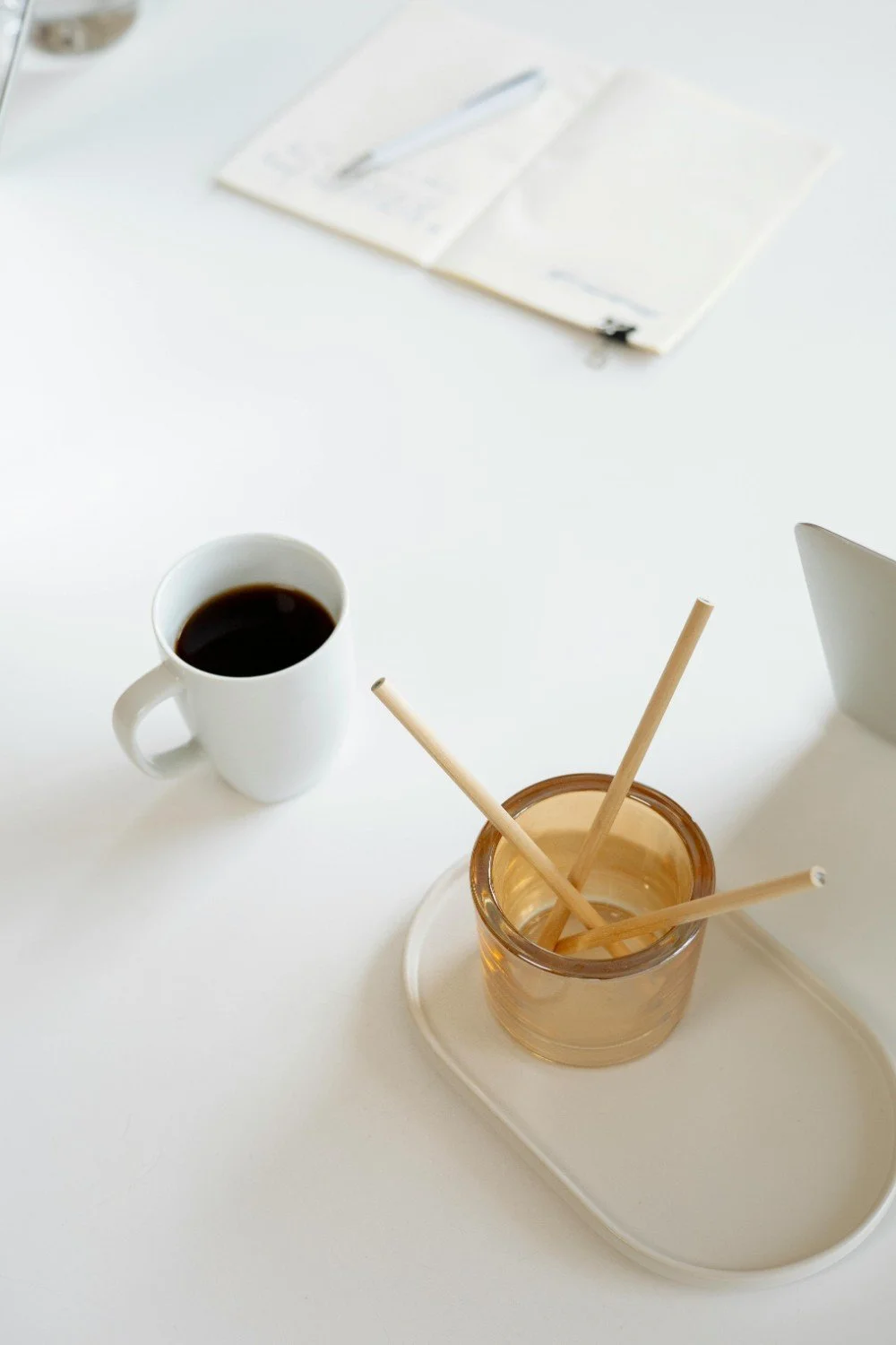White table with a white mug of black coffee, a brown container with wooden stir sticks, and an open notebook with a pen.