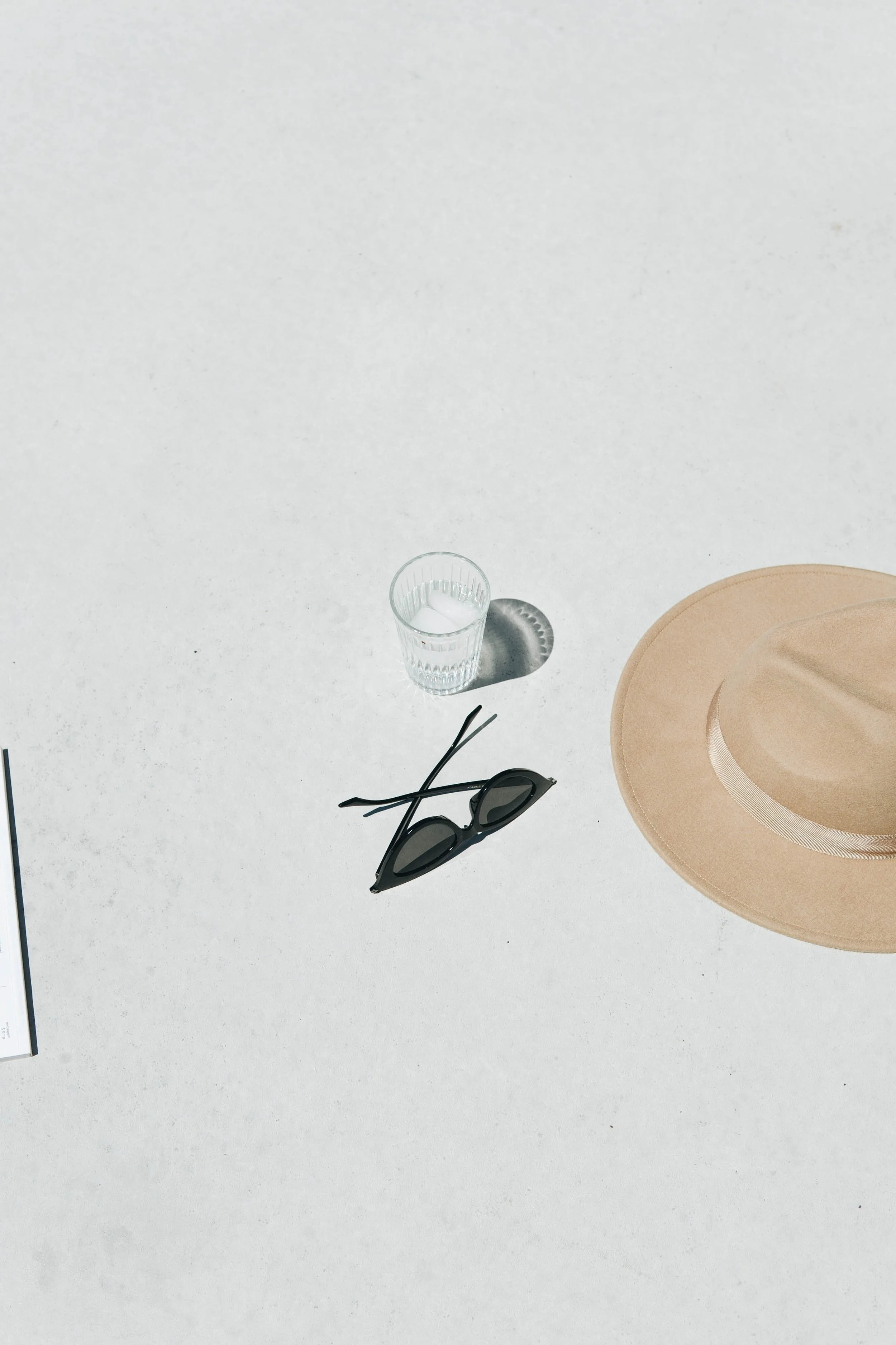 A beige hat, black sunglasses, a glass of water, and part of a book or paper on a white textured surface.