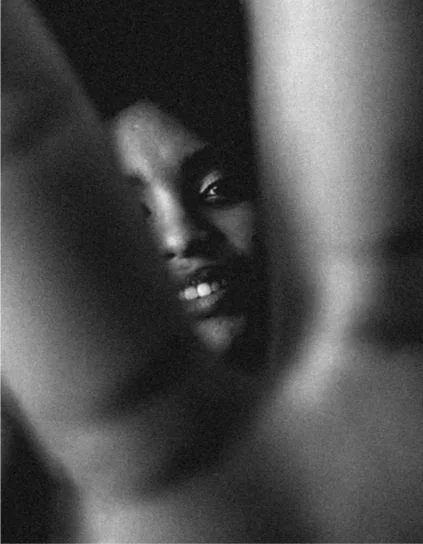 Close-up of a person with dark skin and a partial view of their face, appearing to be behind a blurred object.