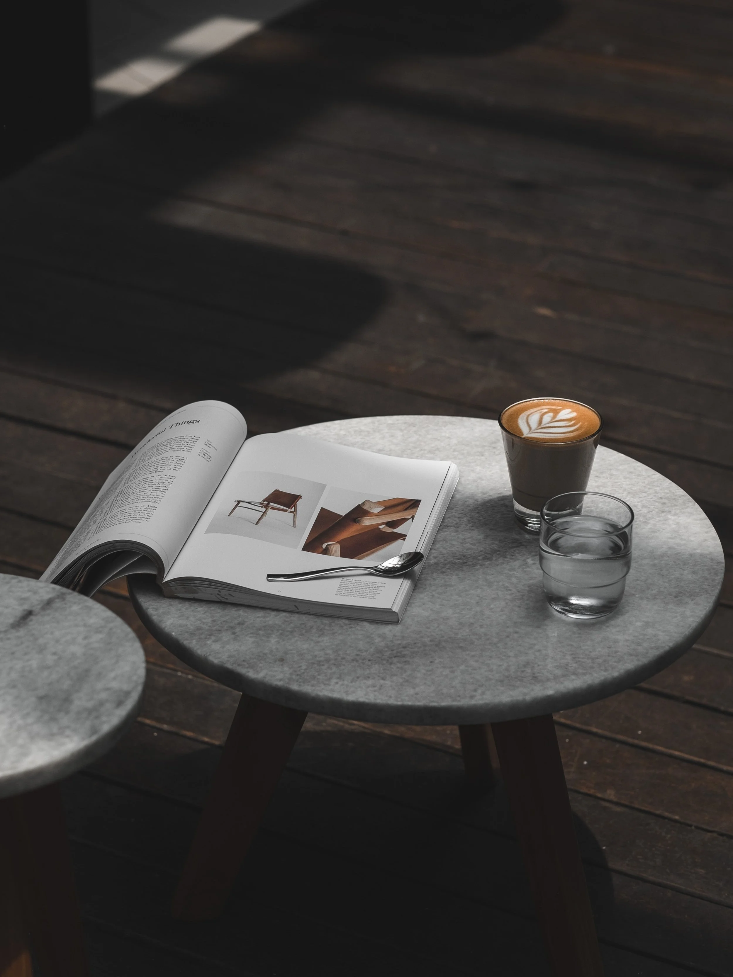 A small round gray table on a wooden floor holds an open magazine, a spoon, a glass of water, and a cup of coffee with latte art.