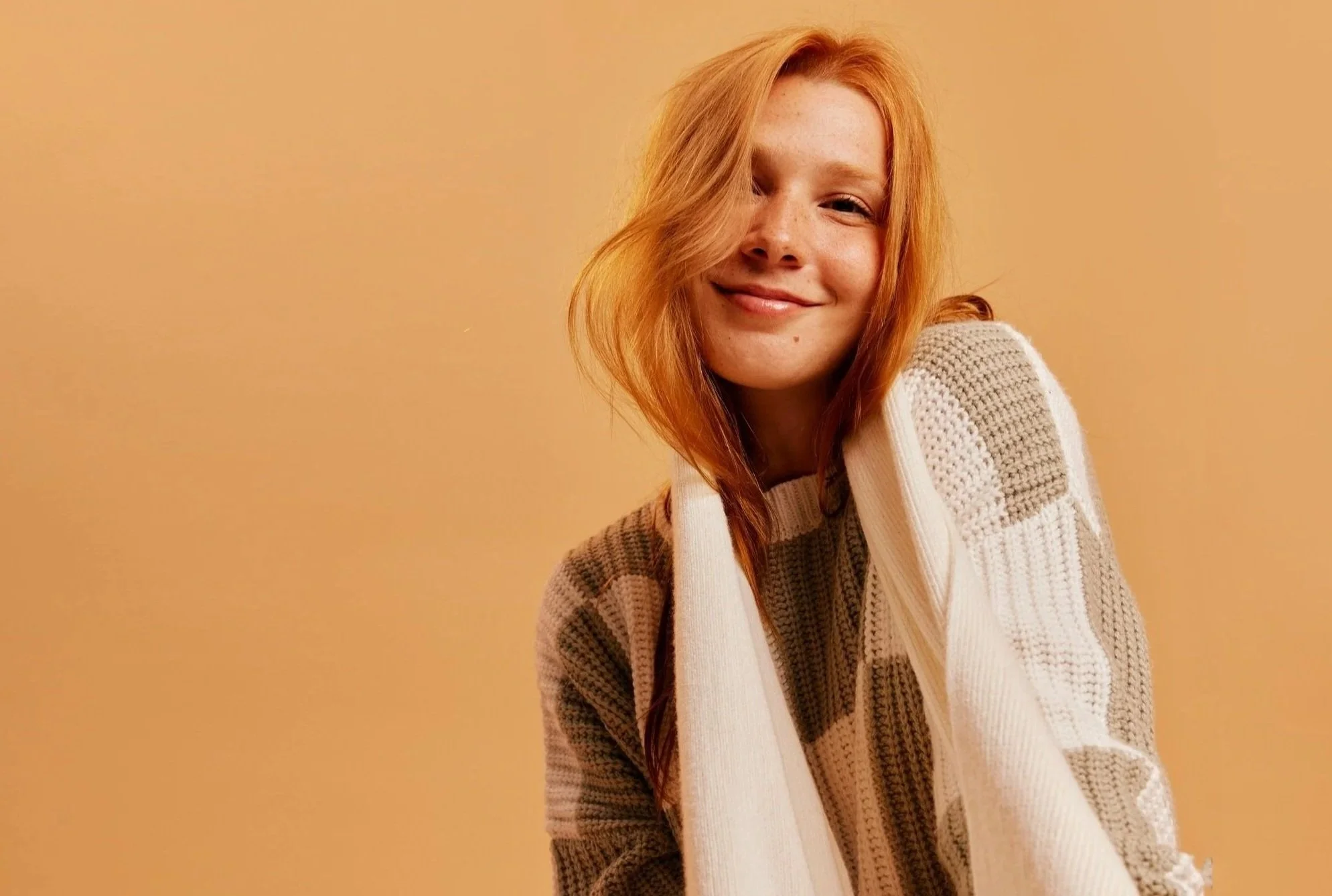 A young woman with long red hair smiling in front of a plain beige background, wearing a beige and white knitted sweater.