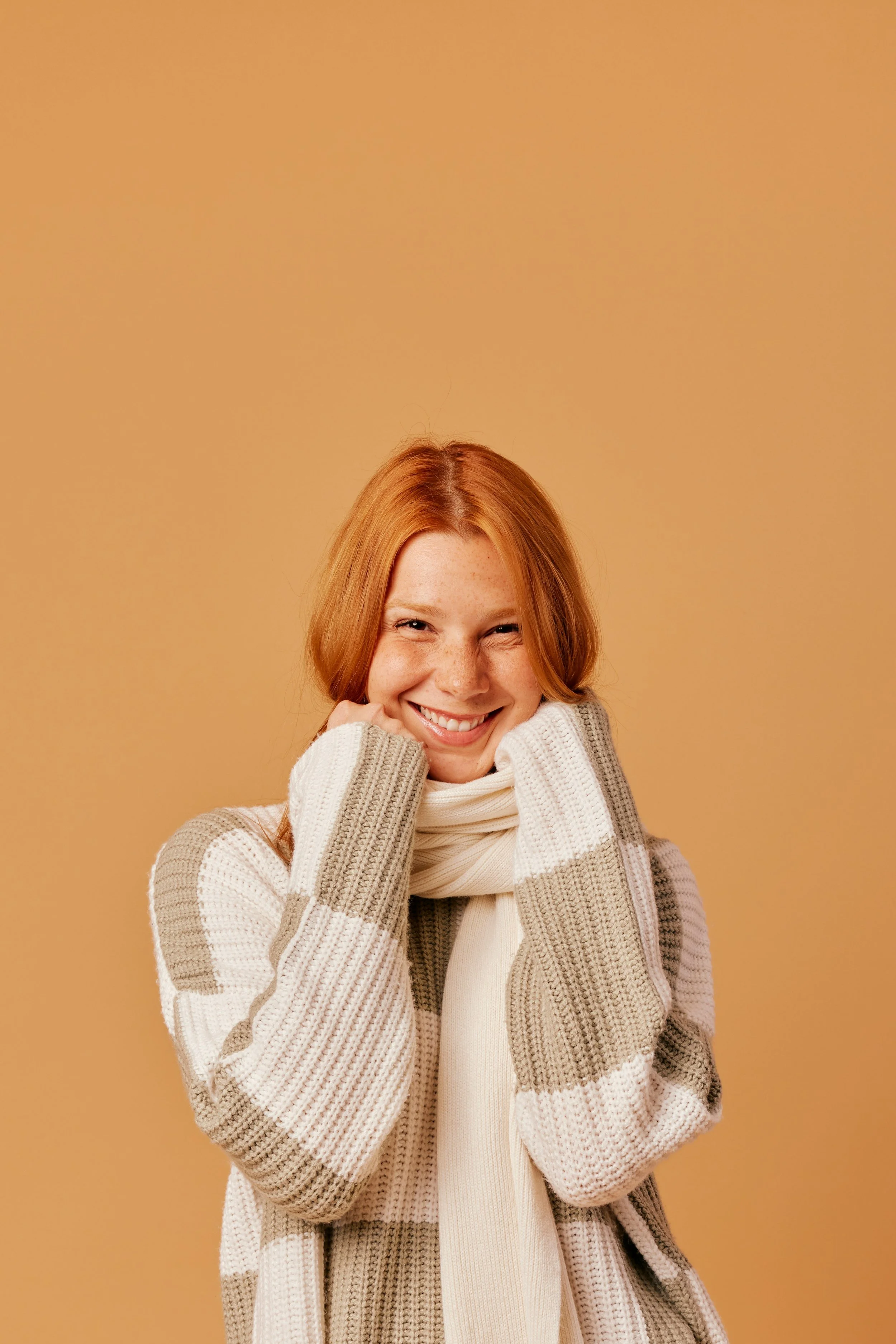 Young woman with red hair smiling, wearing a beige and white striped sweater and a cream scarf, against a plain tan background.